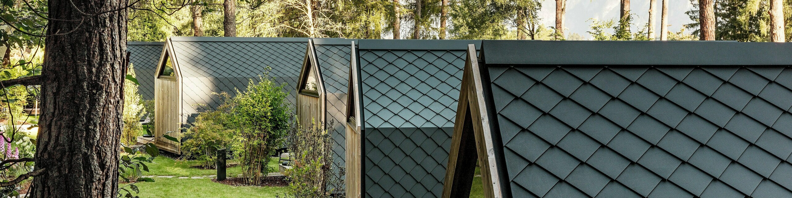 Niekoľko minimalistických minidomov Secret Koi Garden sa harmonicky tiahne pozdĺž zelenej záhradnej osi pri jazere Natterer See. Strechy a fasády sú kompletne obložené drobnými hliníkovými strešnými a fasádnymi šablónami PREFA v odtieni P.10 antracitová, ktoré vytvárajú rovnomerný povrch pripomínajúci rybie šupiny. Obklopené stromami a kríkmi spájajú odolné a poveternostne odolné hliníkové produkty PREFA precíznu architektúru s pokojom, prírodou a kvalitným glampingovým dizajnom.