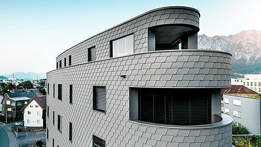 Detailný pohľad na Bernerhaus v Buchsi s jemne zaoblenými rohmi a priebežnými balkónovými pásmi. Fasáda z PREFA fasádnych šindľov v odtieni P.10 svetlošedá pokrýva trojuholníkový priestor ako precízne spracovaná hliníková koža a vytvára živý vzor pripomínajúci šupiny. Homogénny povrch zdôrazňuje plynulé polomery a hru svetla a tieňa. Vďaka ľahkému, odolnému voči poveternostným vplyvom a trvácnemu hliníku PREFA vzniká obvodový plášť budovy s nízkymi nárokmi na údržbu, ktorý má výrazný efekt z diaľky na pozadí alpského prostredia.