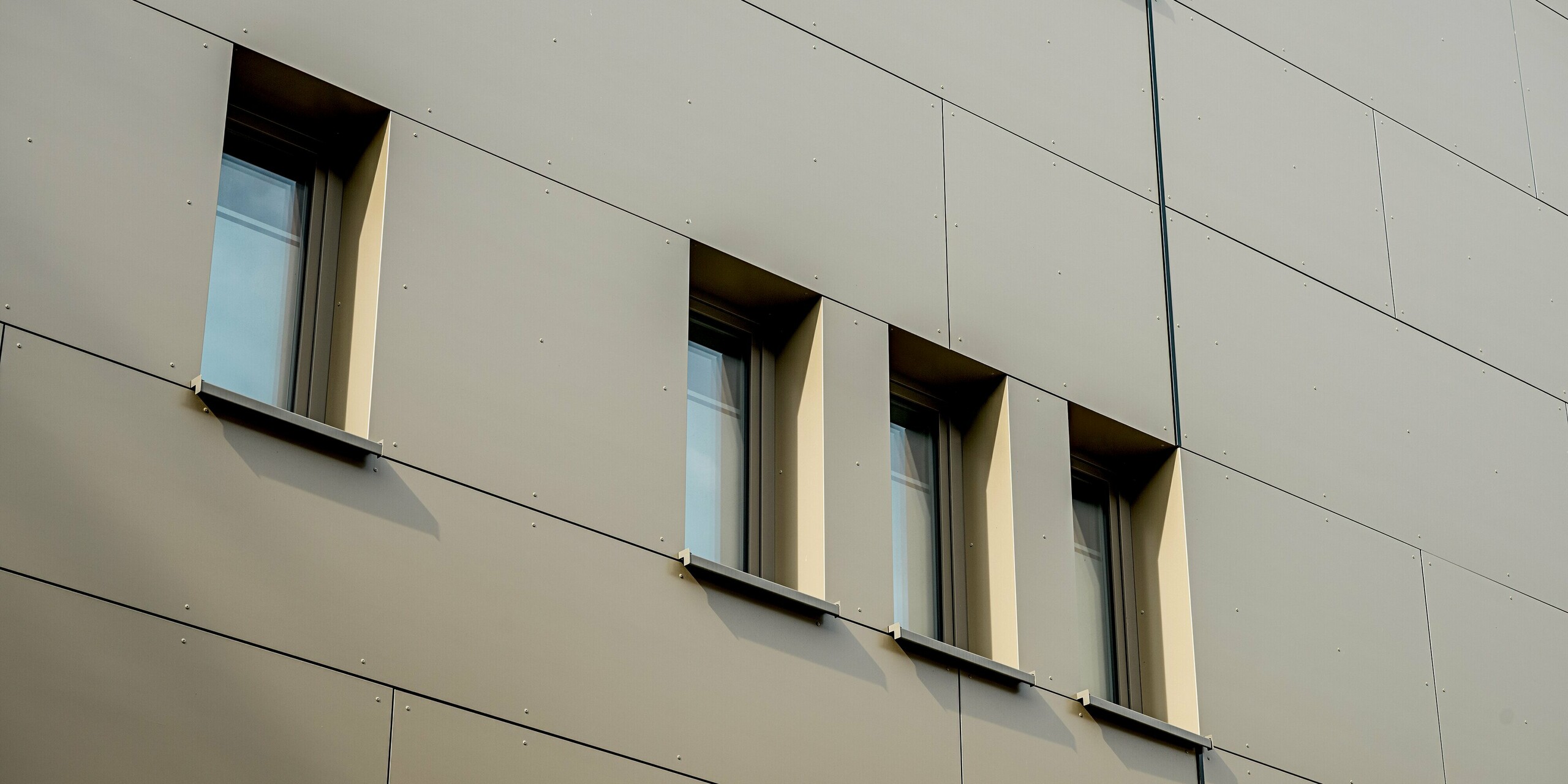 Das Bild zeigt eine Detailaufnahme der Fassade des Festo-Gebäudes in Langenhagen. Hier kommen die eleganten PREFABOND Aluminium Verbundplatten zum Einsatz, die in einem sanften Bronze gehalten sind und der Wand eine ruhige, moderne Ästhetik verleihen. Die Fenster sind in speziell geformte Aussparungen eingefasst, was den minimalistischen Charakter des Gebäudes unterstreicht. Die präzise Anordnung der Fenster und die feine Linienführung betonen das klare Design. Dank der hochfesten und witterungsbeständigen Materialien bleibt die Fassade über Jahre hinweg ästhetisch und funktional, perfekt für langlebige Architektur.