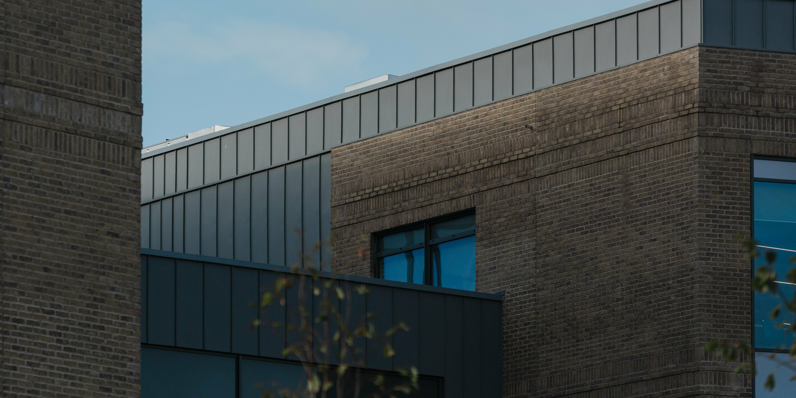 Ein präziser Fassadendetailblick auf das Northern Regional College in Ballymena zeigt den oberen Gebäudeabschluss mit vertikal verlegten PREFALZ Aluminium-Stehfalzbahnen in Patinagrau. Die klaren Linien des Aluminiumbandes kontrastieren wirkungsvoll mit dem warmen Klinkermauerwerk und den tief eingeschnittenen Fensteröffnungen. Die ruhige Metalloberfläche reflektiert das Tageslicht subtil vor blauem Himmel. PREFA Aluminium überzeugt hier durch Langlebigkeit, formale Präzision und eine zeitlose, wartungsarme Lösung für moderne Bildungsbauten.