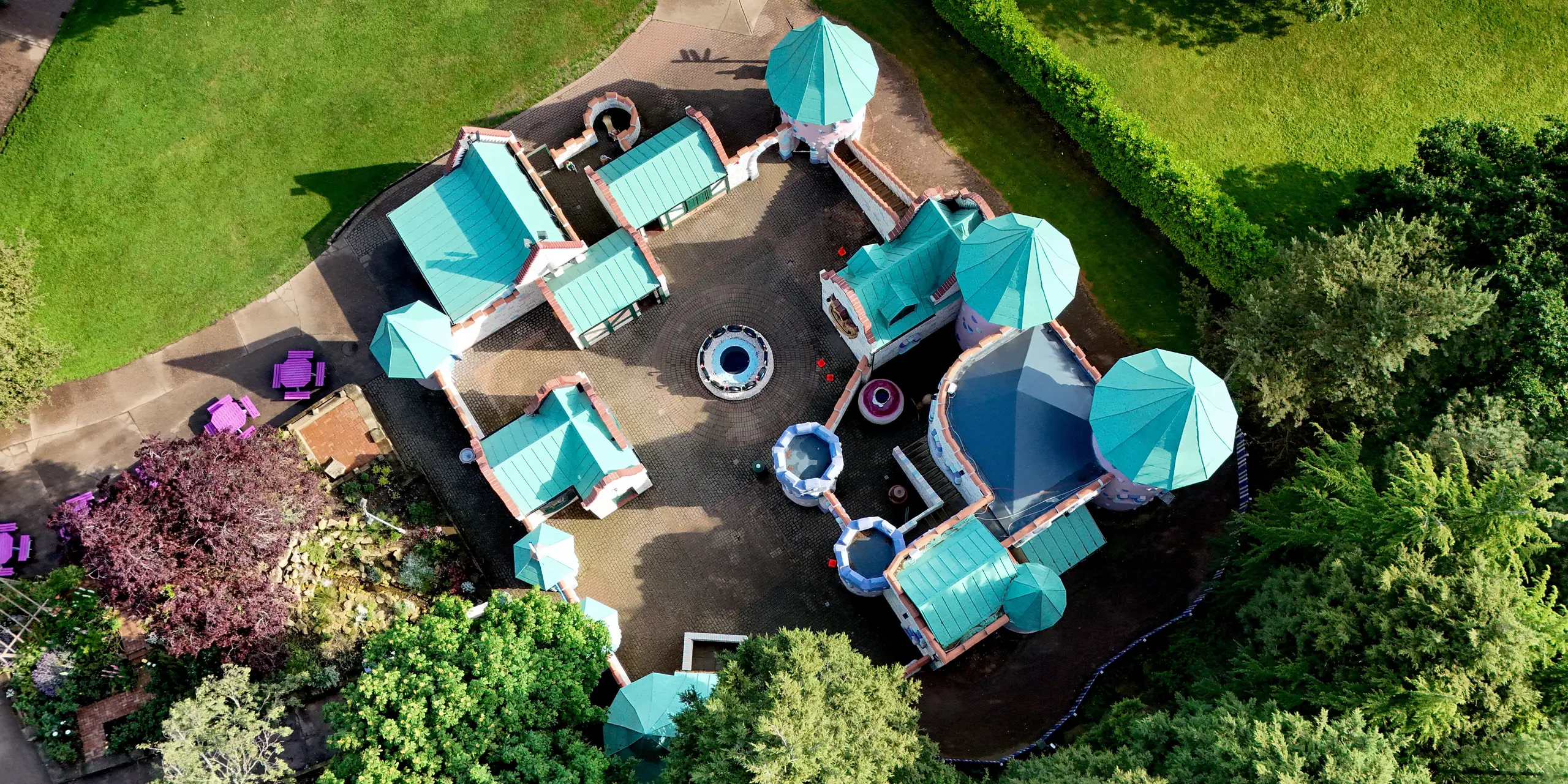 Drohnenaufnahme der märchenhaften Schlossanlage im Sundown Adventureland in Nottinghamshire mit mehreren verspielten Türmen, verwinkelten Grundrissen und markanten, konischen Dächern. Die PREFALZ Aluminiumdächer in P.10 Patinagrün wirken als prägnante Eyecatcher und harmonieren mit den pastellfarbenen Fassaden sowie der grünen Parklandschaft. Die klaren Stehfalze betonen die komplexe Geometrie und zeigen die hohe Formbarkeit des Materials. Gleichzeitig überzeugen die PREFA Dächer durch Langlebigkeit, Korrosionsbeständigkeit und zuverlässigen Schutz bei unterschiedlichsten Wetterbedingungen.