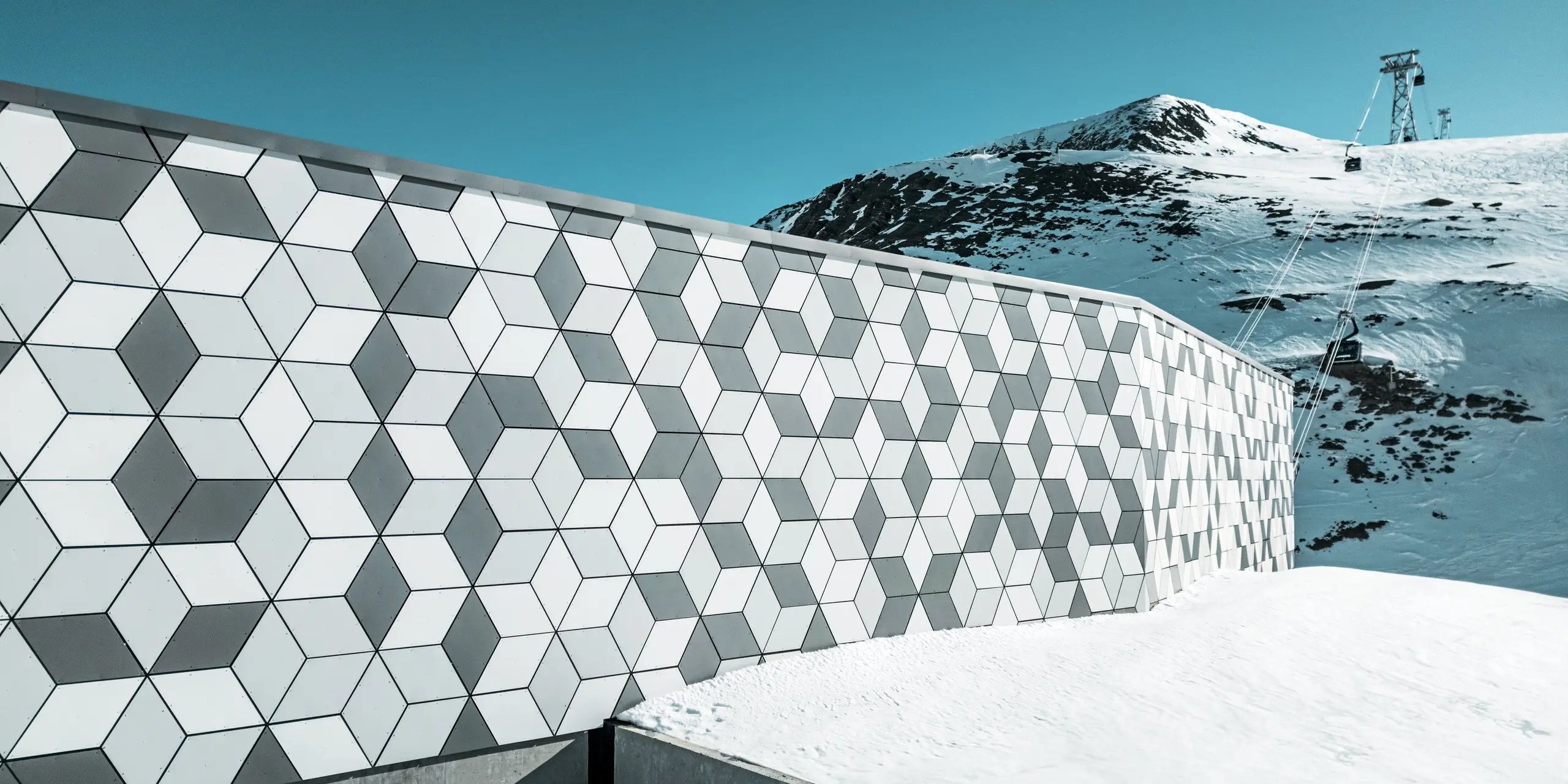 Fasáda stanice lanovky v Les Deux Alpes je majstrovským dielom modernej architektúry. Pozostáva z hliníkových kompozitných panelov PREFABOND usporiadaných do pútavého kosoštvorcového vzoru bielej a sivej farby. Geometrický dizajn harmonicky zapadá do zasneženej horskej krajiny a vďaka robustným materiálom odolným voči poveternostným vplyvom poskytuje dlhodobú ochranu pred extrémnymi poveternostnými podmienkami. Pohľad za fasádou zobrazuje lanovku, ktorá premáva na horu.