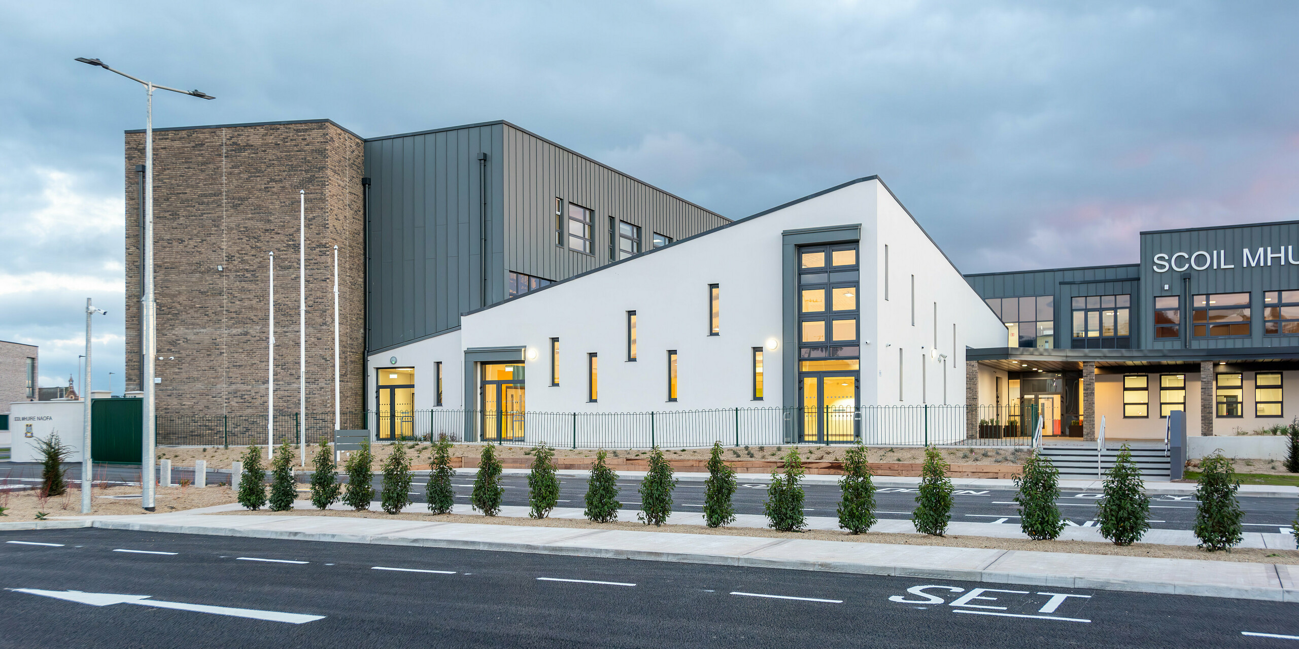 Die moderne Scoil Mhuire Naofa Schule in Carrigtwohill, Irland, mit ihrer eleganten PREFALZ Aluminiumfassade in P.10 Hellgrau. Das helle, minimalistische Design kombiniert klare Linien mit einer hochwertigen Stehfalzdeckung, während die umgebende Landschaft und die neue Zufahrt den modernen Charakter der Bildungseinrichtung unterstreichen. Die beleuchteten Innenräume lenken den Blick auf die großzügigen Fenster.