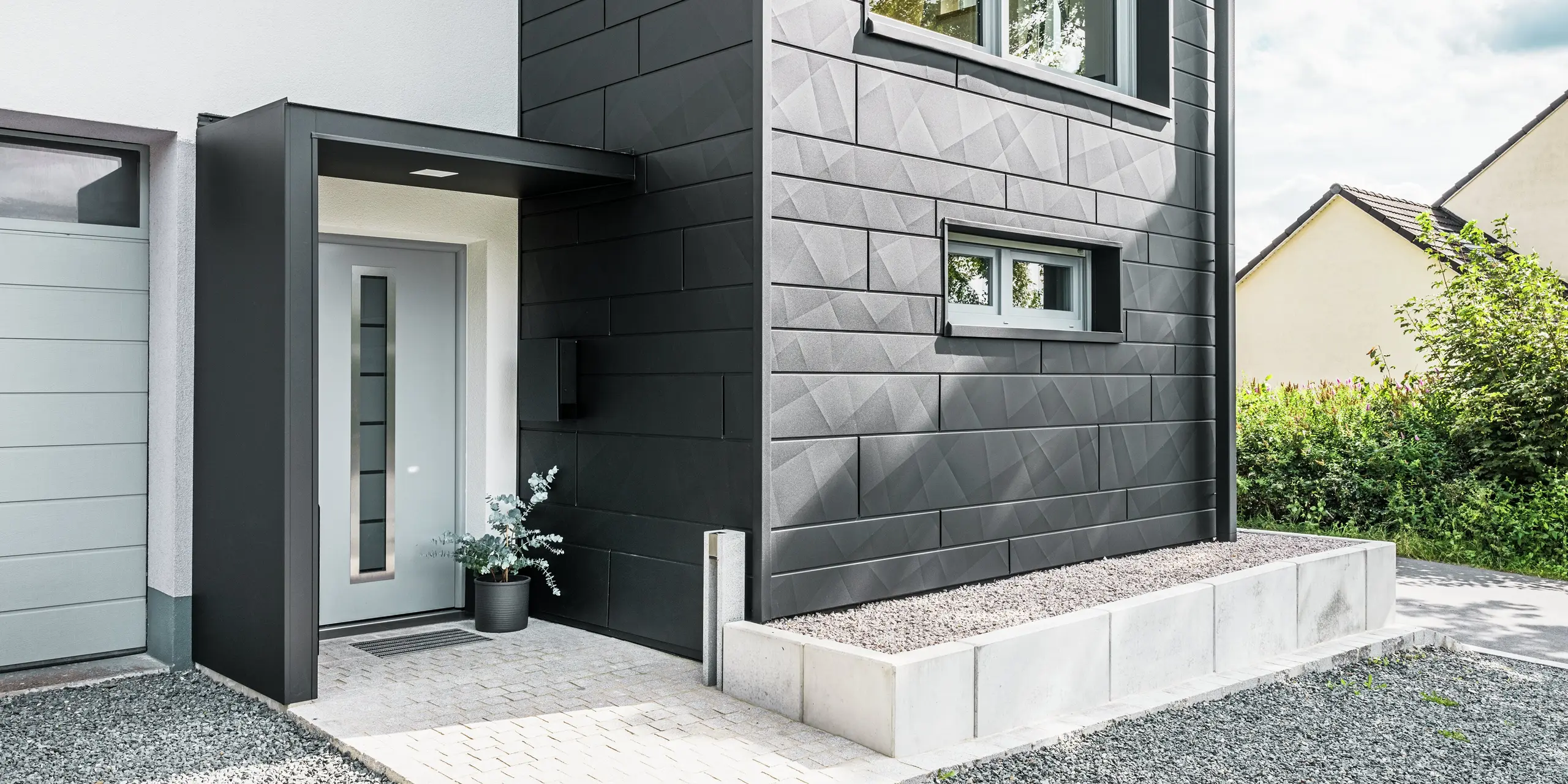Elegantná fasáda PREFA Siding.X v čiernej farbe P.10 dominuje vstupnej časti zmodernizovaného rodinného domu v Teuschnitzi. Vysoko kvalitná plechová fasáda dodáva budove moderný a nadčasový charakter. Jasne štruktúrované fasádne panely vytvárajú výrazný geometrický dizajn, zatiaľ čo čierne hliníkové kompozitné dosky rámujú vchodové dvere a okná. Hranatý zvod PREFA sa bezproblémovo začleňuje do náročného dizajnu a podčiarkuje funkčný a estetický účinok renovácie. Cesta k domu je pokrytá dlažobnými kockami a štrkom.