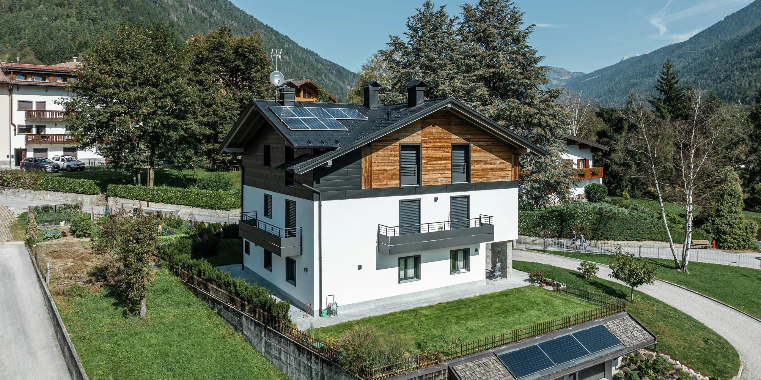 Modernizovaný obytný dom v Pelugo, Taliansko, so strešným panelom PREFA R.16 vo farbe P.10 antracit a fasádou PREFA Siding.X. Kombinácia hliníka, dreva a omietky zabezpečuje odolnú, poveternostným vplyvom odolnú a štýlovú architektúru v alpskom prostredí. Solárne panely na streche a garáži dopĺňajú koncept udržateľnej energie.
