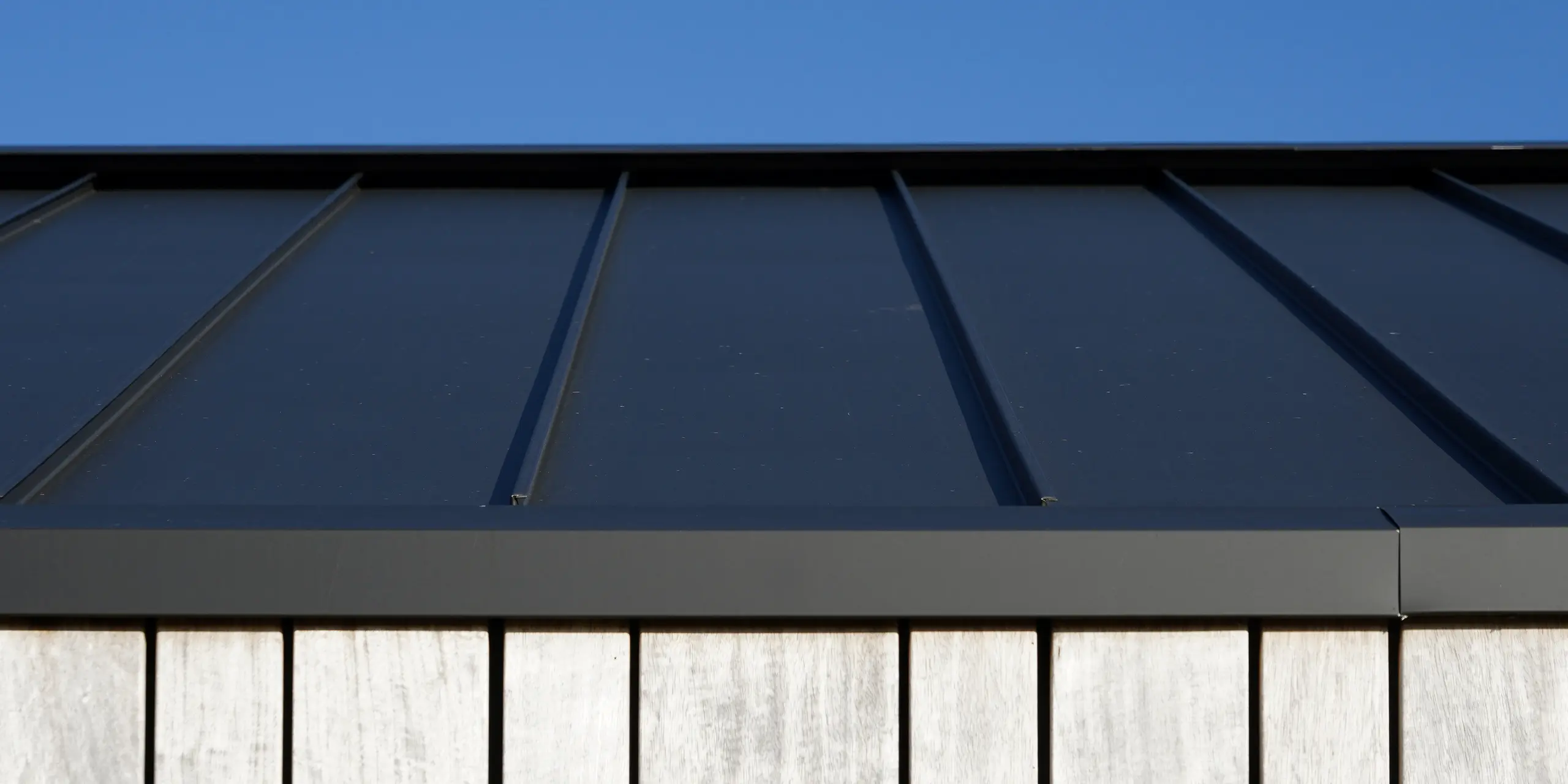 Detailný záber na moderný rodinný dom v holandskom meste Uithoorn so strešnou krytinou PREFALZ so šikmou stojatou drážkou v čiernosivej farbe. Na obrázku je vidieť precízne spracovanie vysokokvalitného plechu PREFA v kombinácii s prírodnými drevenými prvkami na fasáde. Jasné línie robustnej hliníkovej strechy podčiarkujú trvanlivú konštrukciu odolnú voči poveternostným vplyvom.