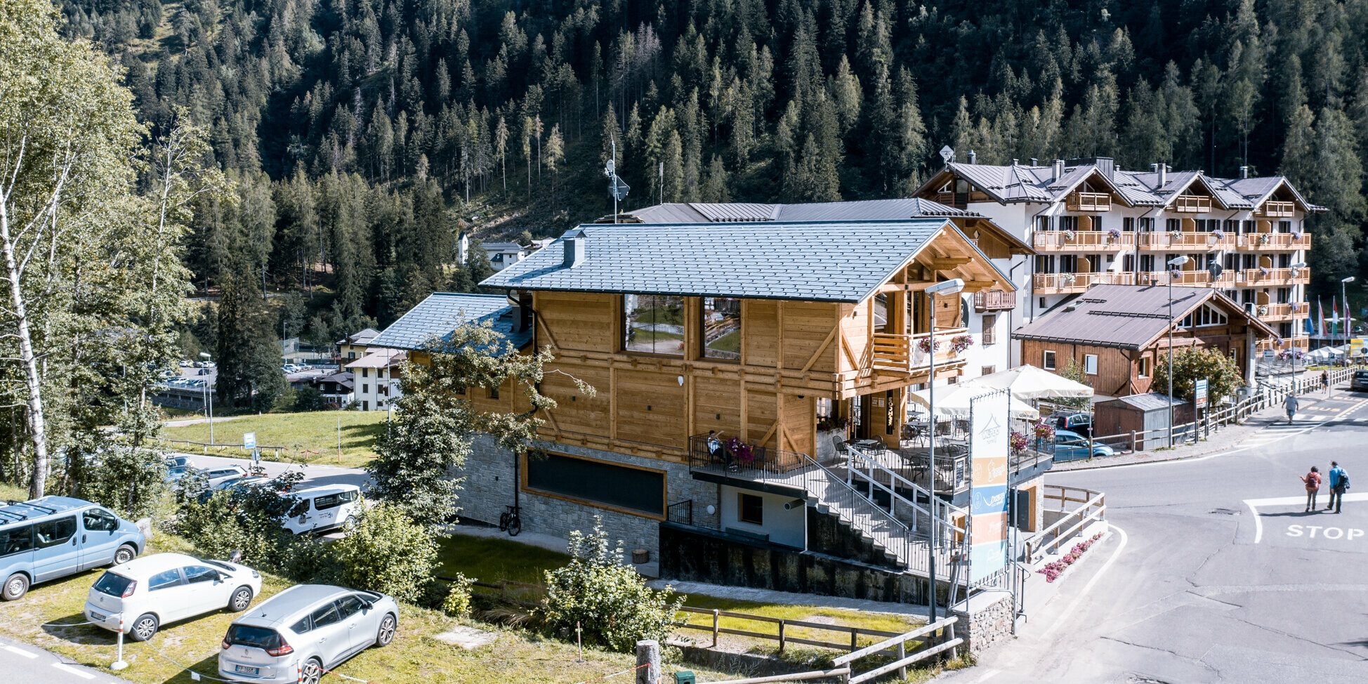 Letecký pohľad na alpský apres-ski bar v Taliansku, ktorý je zvýraznený odolným hliníkovým strešným systémom. Robustné strešné šindle PREFA DS.19 vo farbe P.10 kamenná šedá chránia budovu pred nepriaznivými vplyvmi počasia. Kontrastná kombinácia drevenej konštrukcie a kamenných sivých akcentov, obklopená hustými borovicovými lesmi a tradičnou chatovou architektúrou, vytvára pre návštevníkov príjemnú atmosféru a harmonicky zapadá do prírodného prostredia.