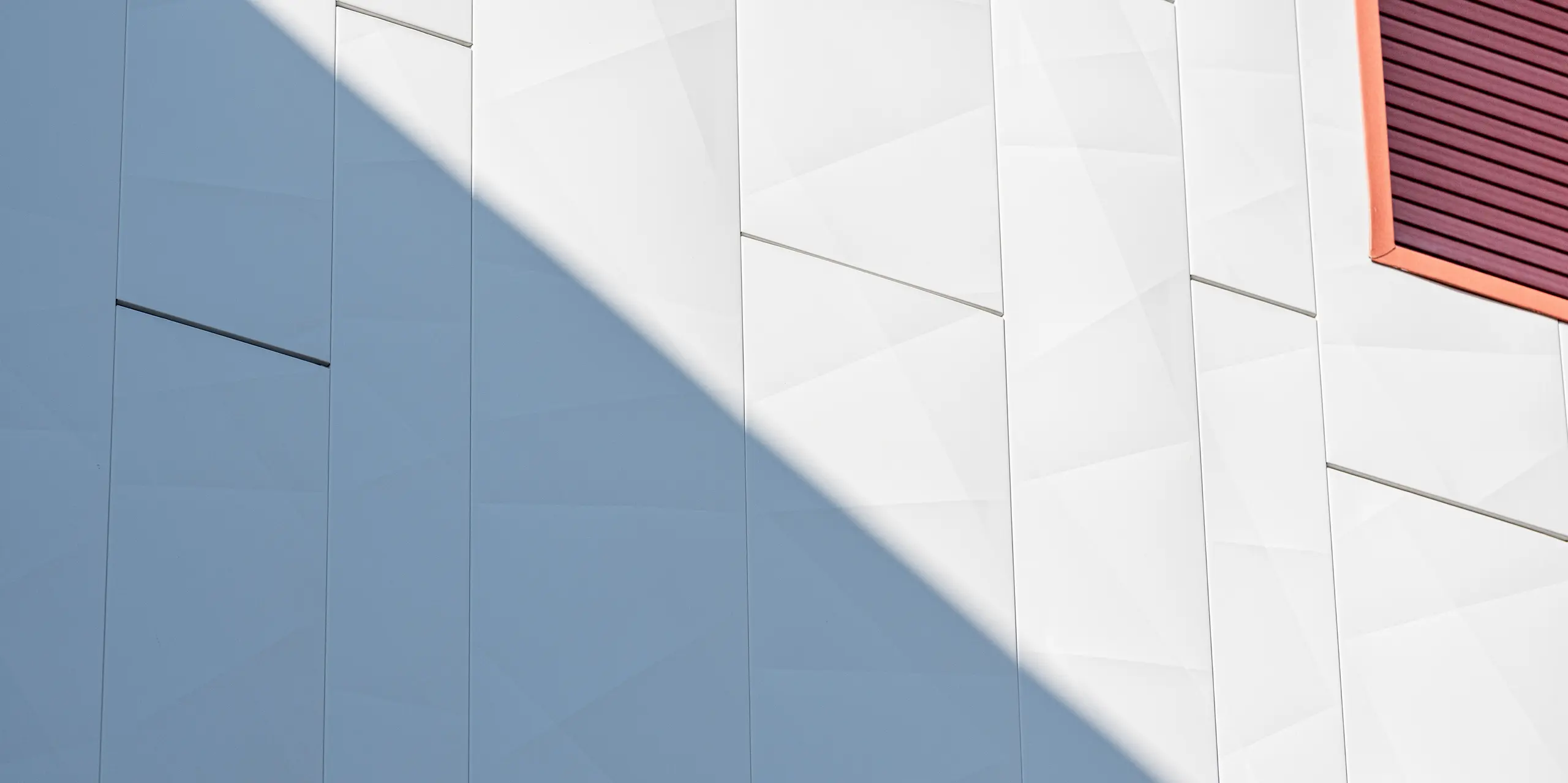 Detailaufnahme der sanierten Fassade der Wohnanlage Alba B mit PREFA Siding.X Aluminiumfassadenpaneelen: Großformatige, vertikal verlegte Paneele mit feinen Kantungen erzeugen eine lebendige, geometrische Struktur. Ein schräg einfallender Schatten betont die metallische Oberfläche und sorgt für eindrucksvolle Licht- und Schattenspiele. Am rechten Bildrand setzt eine rot akzentuierte Fensterlaibung einen starken Kontrast. Die PREFA Aluminiumfassade überzeugt durch hohe Widerstandsfähigkeit, Langlebigkeit und eine dauerhaft wartungsarme, ästhetische Gebäudehülle.