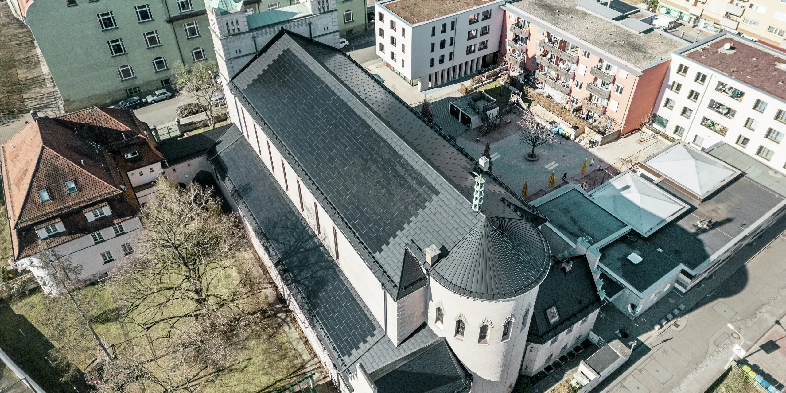 Pohľad z vtáčej perspektívy na zadnú časť kostola svätého Karola Boromejského v Norimbergu. Čierne integrované solárne panely PREFA pokrývajú nápadnú okrúhlu šikmú strechu nad hlavnou časťou kostola, ako aj strechu prístavby. Hliníkové strešné panely s integrovanými fotovoltickými modulmi dokonale ladia s historickým vzhľadom kostola. Pohľad na moderné okolie so susednými obytnými budovami a zelenou plochou nádvoria ilustruje úspešnú kombináciu ochrany pamiatky, modernej architektúry a udržateľnej výroby energie. Ideálne riešenie na ochranu pamiatok v mestskom prostredí.