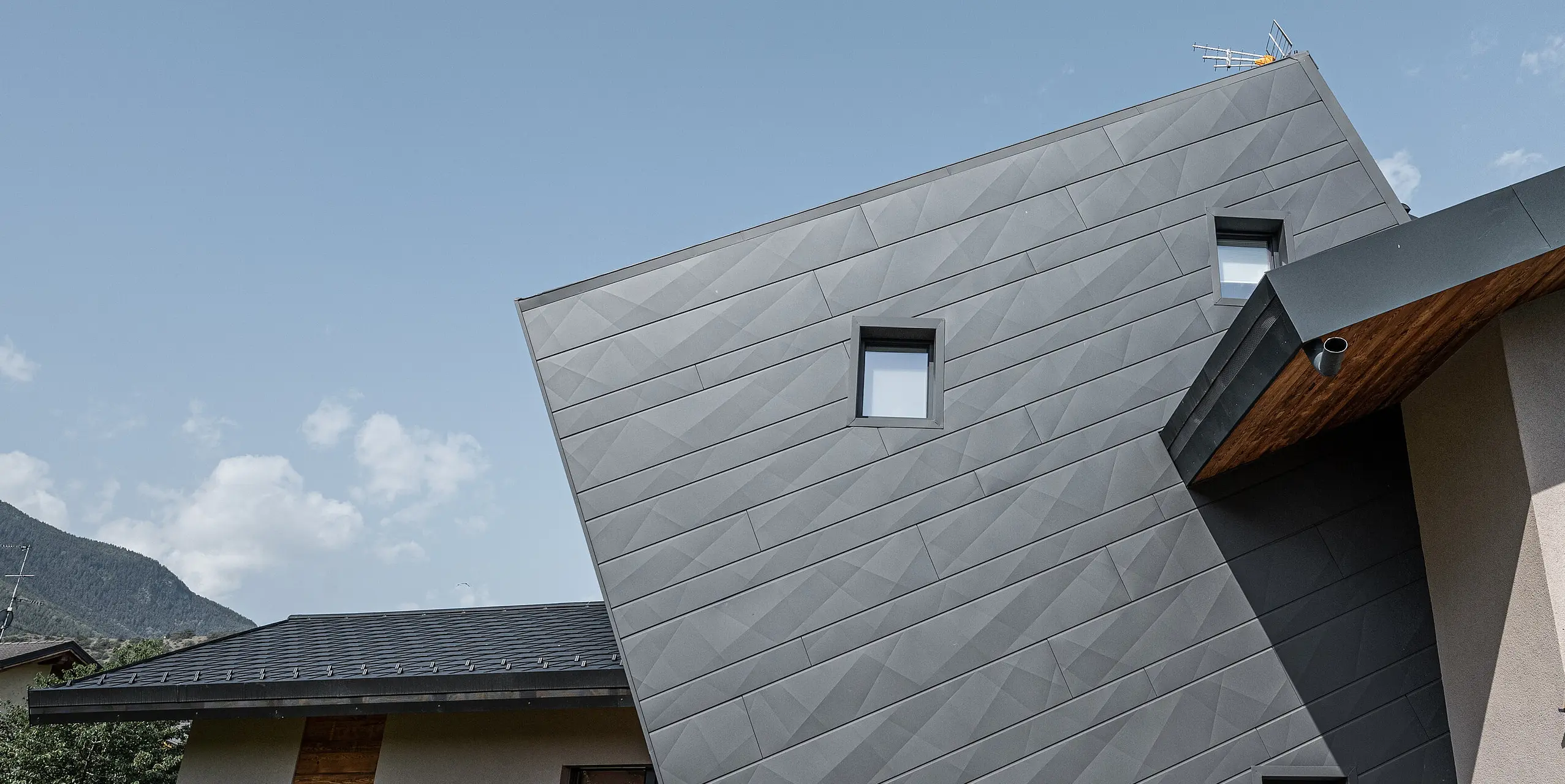 Vila Tia v talianskom Gressane má moderný dizajn fasády s obkladom PREFA Siding.X vo farbe P.10 tmavošedá. Dynamické usporiadanie panelov vytvára trojrozmerný vzhľad, ktorý je zdôraznený kontrastom s prírodnými drevenými prvkami a okolitou krajinou. Toto inovatívne špeciálne riešenie šikmých fasádnych panelov Siding.X je symbolom moderného a odolného dizajnu.