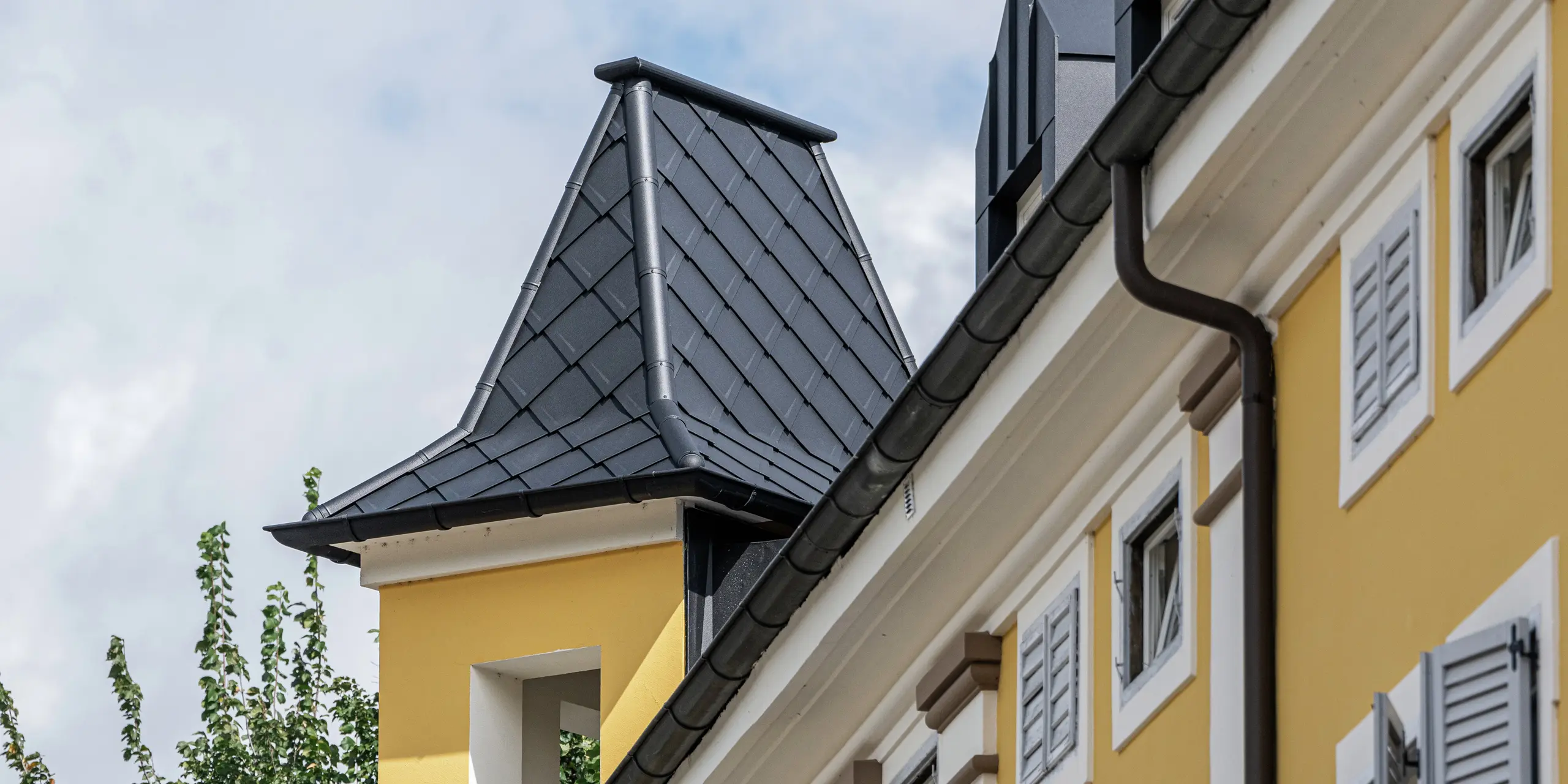 Detailný pohľad na historickú vežu hotela Jarolim v Brixene, navrhnutú s použitím hliníkových strešných šindľov PREFA. Odolné voči poveternostným vplyvom a ľahké produkty PREFA zachovávajú historický charakter budovy a zabezpečujú trvalé a estetické riešenie strechy. Vysokokvalitné spracovanie vikierov a obloženia veže podčiarkuje spojenie tradičnej architektúry a modernej techniky – ideálne pre pamiatkovo chránené budovy.
