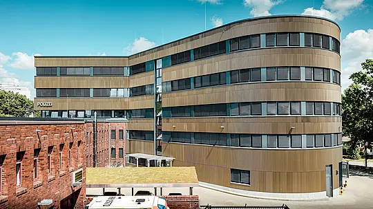 Dynamický pohľad na exteriér rozšíreného policajného komisariátu č. 43 v Hamburgu: Zakrivená nová budova s hliníkovou fasádou PREFA o rozlohe približne 1 100 m², tvorenou eloxovanými extrudovanými profilmi „Zacke“ v odtieni strednej bronzy, púta pozornosť a vytvára výrazný kontrast k existujúcej tehlovej budove. Jemne štruktúrovaný povrch živým spôsobom hrá so svetlom a tieňmi a dodáva fasáde hĺbku a precíznosť. Vďaka vysokej odolnosti proti korózii, dlhej životnosti a nízkym nárokom na údržbu predstavujú hliníkové riešenia PREFA udržateľnú, funkčnú a architektonicky náročnú kvalitu stavby.