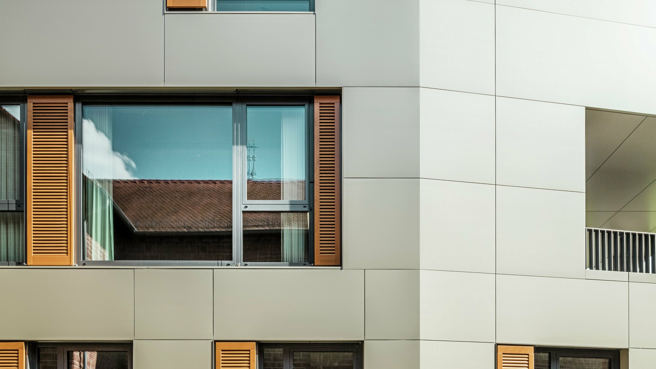 Detailný záber fasády radnice v Alzenau v Nemecku s hliníkovými kompozitnými doskami PREFA v bronzovej farbe. Veľké okná sú orámované elegantnými drevenými žalúziami v teplej hnedej farbe, ktoré dodávajú fasáde príjemnú textúru. V popredí je vidieť upravený kvetinový záhon, ktorý dopĺňa mestský dizajn budovy zelenými akcentmi. Jasná obloha a čisté línie architektúry podčiarkujú moderný charakter budovy.