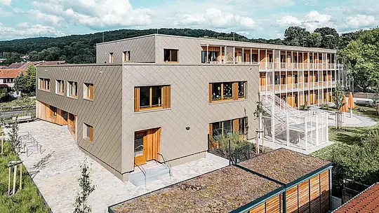 Z vyvýšenej perspektívy zobrazuje snímka novostavbu materskej školy v Alzenau v tvare písmena L s nadväzujúcim obytným traktom – progresívnu hybridnú budovu realizovanú udržateľným spôsobom výstavby. Predsadená bronzová hliníková fasáda PREFA s fasádnymi šablónami 44 × 44 formuje moderný, štruktúrovaný vonkajší plášť časti určenej pre materskú školu. Veľkorysé drevené okná a vertikálne lamely vytvárajú prívetivý a teplý vzhľad. Zadná časť s vonkajším schodiskom, hernými plochami a zelenými plochami poukazuje na funkčné oddelenie a zároveň prepojenie jednotlivých spôsobov využitia. Zelená strecha spolu s kombináciou hliníka, dreva a betónu odrážajú vysoké ekologické ambície projektu – silné vyhlásenie v prospech udržateľnej architektúry vzdelávacích a obytných stavieb.