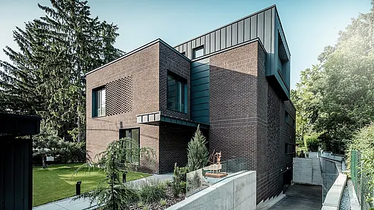Záber zachytáva modernú architektúru budapeštianskej viacgeneračnej vily s výraznou kombináciou tmavočerveného klinkeru a hliníka PREFALZ v odtieni P.10 antracit. Jasne definovaný kubus s vertikálnymi stojatými falcami vytvára silný kontrast k štruktúrovanej tehlovej fasáde pod ním. Elegantne riešený vjazd do podzemnej garáže a starostlivo upravená záhradná architektúra podčiarkujú vysokú kvalitu realizácie. Trvácne a poveternostným vplyvom odolné hliníkové produkty PREFA dodávajú budove nadčasový, precízny a architektonicky výrazný vzhľad.