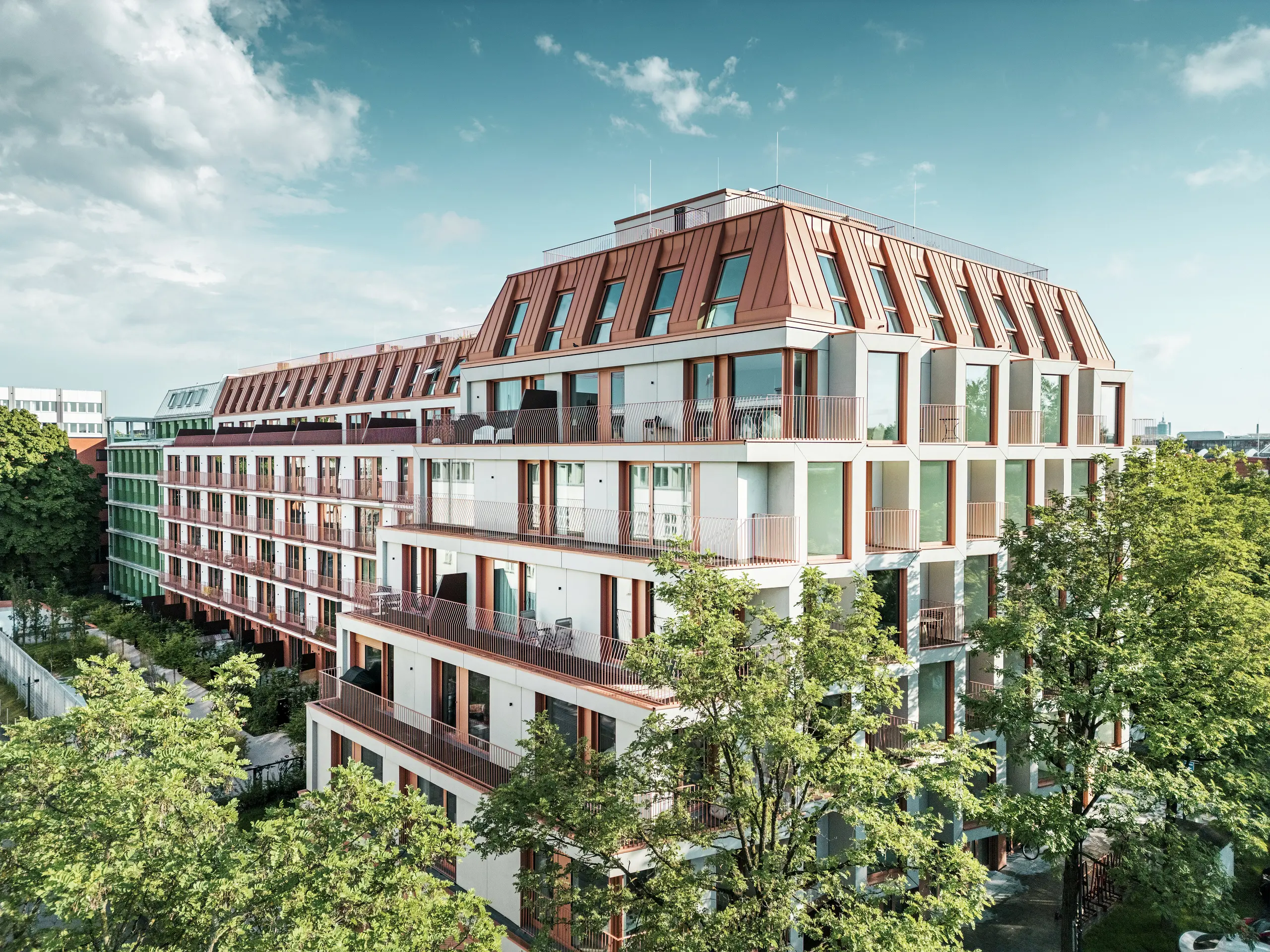Celkový pohľad na mestský obytný projekt Van B v Mníchove s čistou architektúrou, zelenými vonkajšími plochami a výraznou hliníkovou strechou PREFALZ vo farbe IGP Orange HDP od spoločnosti PREFA. Odolný a na údržbu nenáročný strešný materiál tvorí jednotiaci dizajnový prvok budovy a poskytuje dlhodobé a estetické riešenie v mestskom priestore. Veľkorysé okenné plochy, balkóny a premyslené pôdorysy ponúkajú moderný komfort bývania s vysokou kvalitou dizajnu.