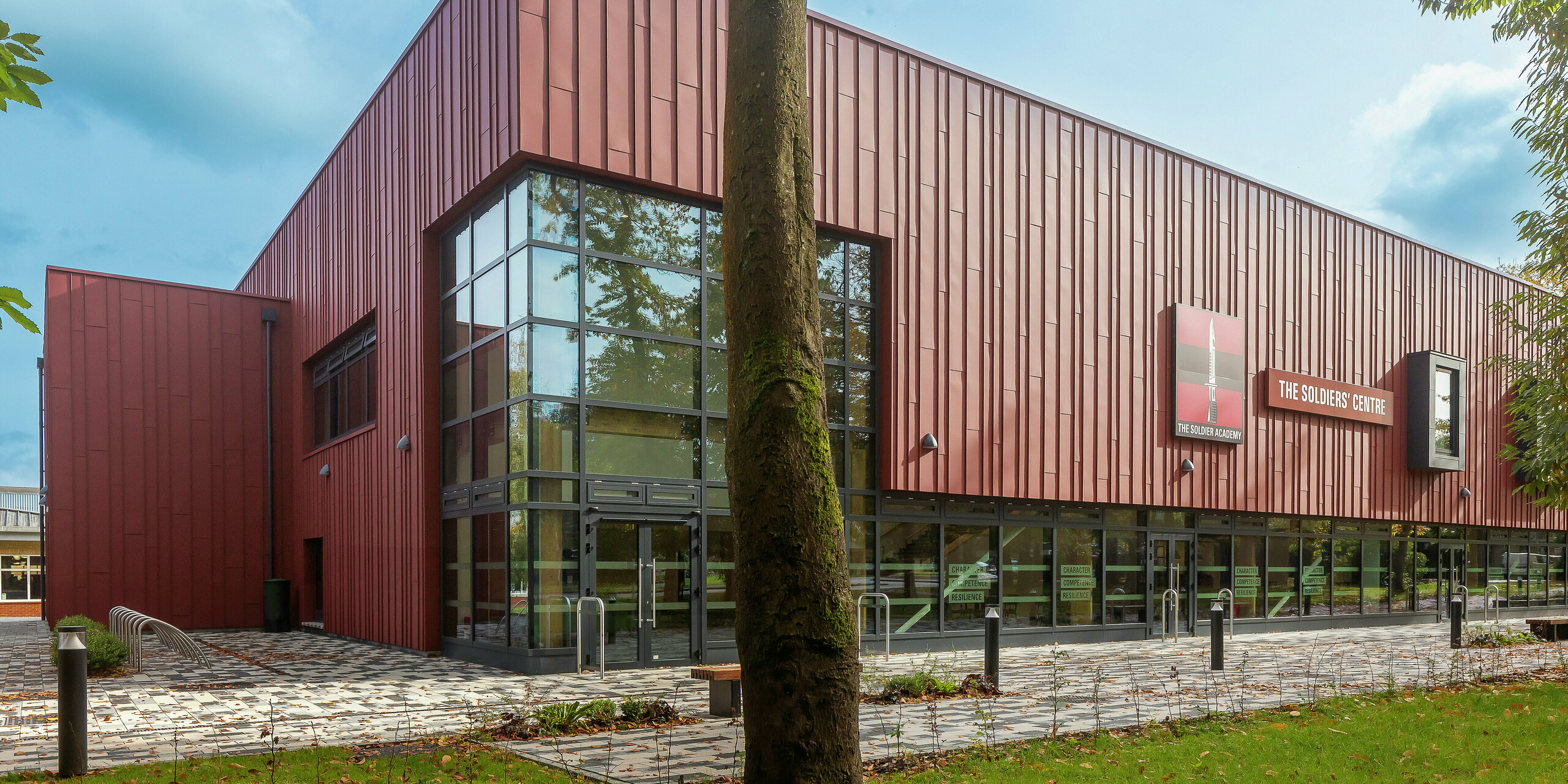Inovatívny dizajn fasády s PREFALZ: Soldiers' Centre v Pirbrightu zaujme moderným obložením zo stojatých falcov PREFALZ v odtieni P.10 tmavočervená. Veľké sklenené plochy zaisťujú svetlú, príjemnú atmosféru, okenné rámy v odtieni P.10 Anthrazit dodávajú štýlové akcenty. Udržateľnosť, dlhá životnosť a dizajn v jednom – vysoko kvalitné hliníkové produkty PREFA pre architektúru budúcnosti