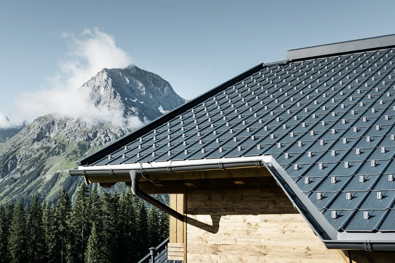 Falcovaná škridla PREFA v alpskom prostredí – falcovaná škridla v antracitovej farbe položená na chate v obci Lech am Arlberg s horami v pozadí; PREFA odvodňovací systém v atracitovej farbe, drevená fasáda