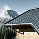Falcovaná škridla PREFA v alpskom prostredí – falcovaná škridla v antracitovej farbe položená na chate v obci Lech am Arlberg s horami v pozadí; PREFA odvodňovací systém v atracitovej farbe, drevená fasáda