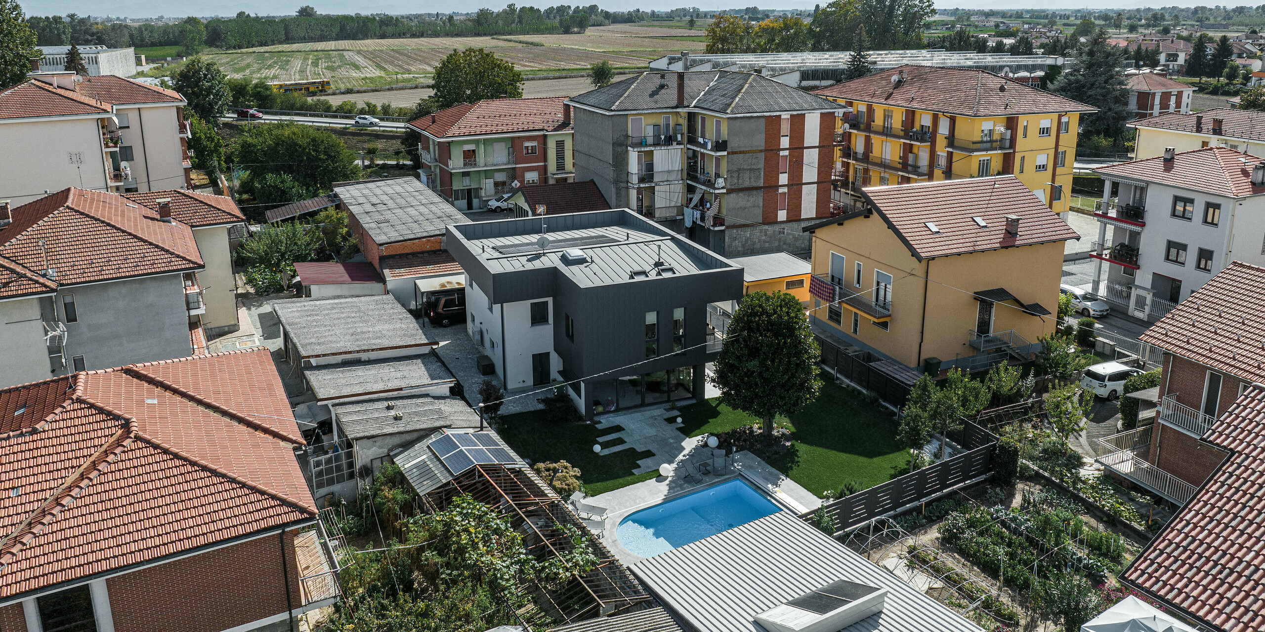 Letecký pohľad na moderný rodinný dom v Savigliane v Taliansku s tmavošedou hliníkovou fasádou PREFA Siding.X. Tento pohľad z vtáčej perspektívy ukazuje dom zasadený do malomestskej štvrte s okolitými tradičnými budovami a jasným výhľadom na vzdialené hory. Elegantná a nenápadná fasáda domu vyniká v pestrom susedstve, zatiaľ čo udržiavaná záhrada s bazénom ponúka oázu oddychu