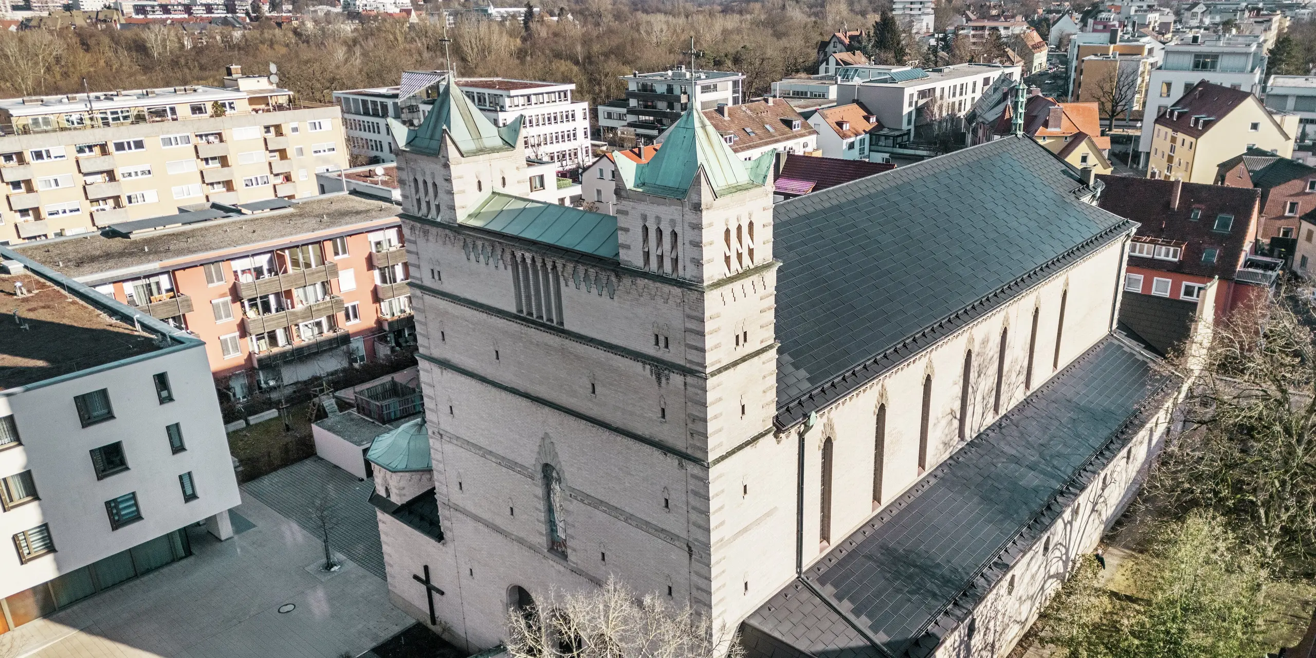 Priečelie kostola svätého Karola Boromejského v Norimbergu: Na obrázku je impozantná strecha pamiatkovo chráneného kostola, ktorá je pokrytá čiernymi integrovanými solárnymi panelmi PREFA. Solárne moduly harmonicky zapadajú do historickej architektúry kostola a zároveň predstavujú moderné riešenie pre udržateľnú výrobu energie. V pozadí je vidieť moderné budovy a zelené kopce Norimbergu, ktoré zdôrazňujú úspešný prechod medzi tradičnou architektúrou kostola a súčasným zásobovaním energiou. Hliníkovú strechu kostola dopĺňa štýlový strešný panel PREFA R.16.