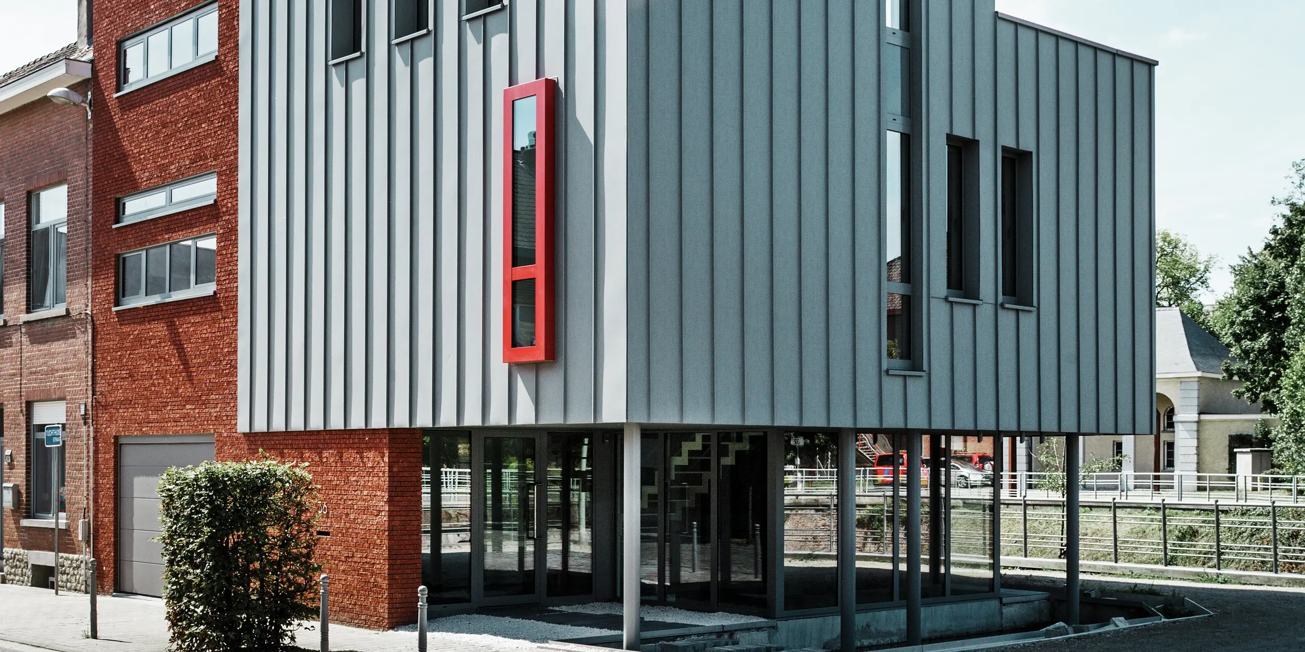 Moderná obytná a administratívna budova vo Vilvoorde v Belgicku s výraznou hliníkovou fasádou PREFALZ v patinovo šedej farbe. Vertikálna konštrukcia so stojatým falcom zaisťuje elegantné línie, zatiaľ čo jasne červené okná pútajú pozornosť. Veľkoplošné zasklenie a prízemie pôsobiace ako vznášajúce sa dodávajú budove ľahkosť a architektonickú rafinovanosť.