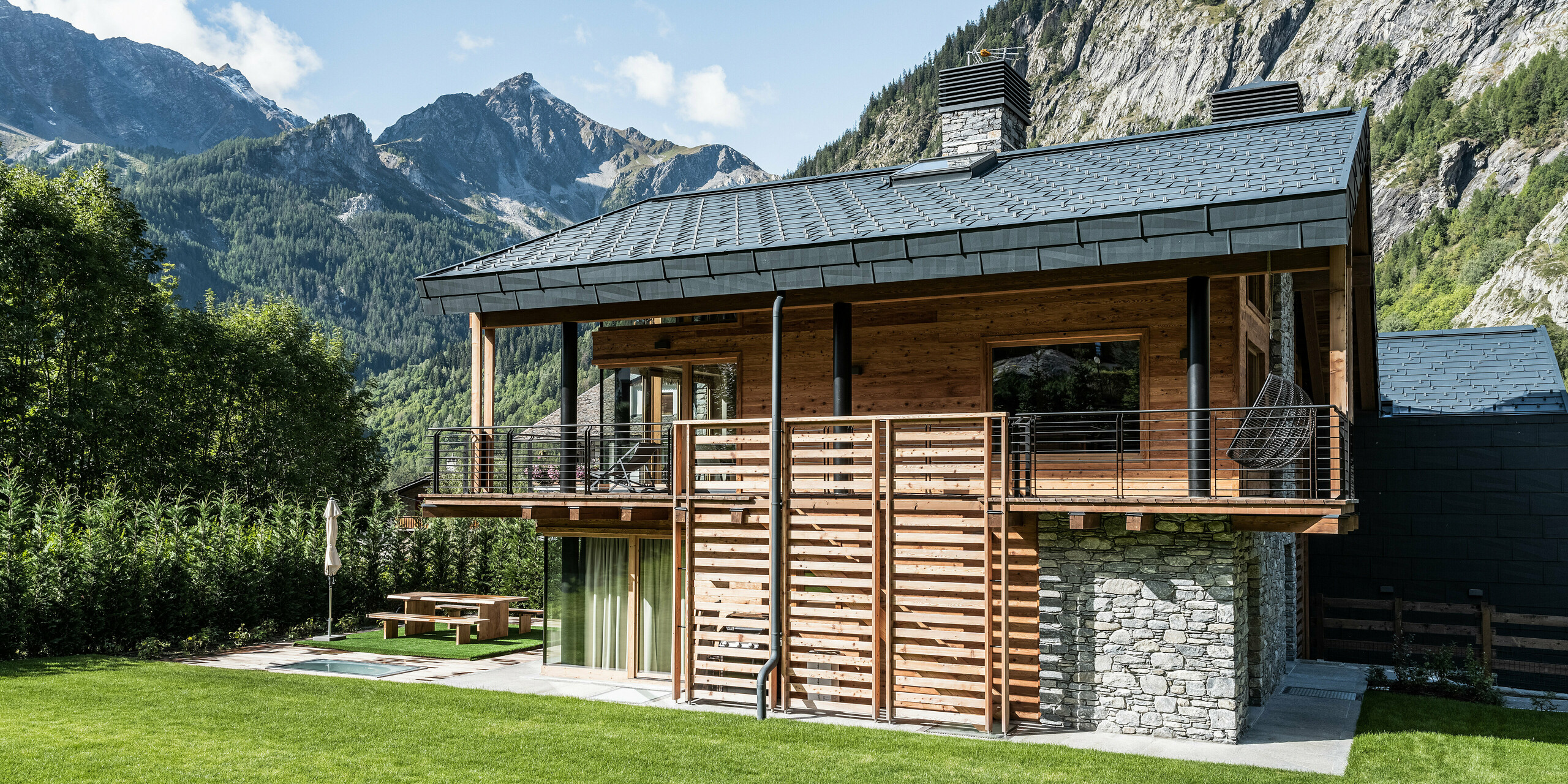 Luxusná vila v Courmayeur s fasádou z prírodného kameňa a dreva a elegantnou strechou z hliníkových panelov PREFA FX.12 vo farbe P.10 kamenná šedá. Priestranné balkóny ponúkajú úchvatný výhľad na okolité hory. Dobre udržiavaná záhrada s posedením a starostlivo upraveným trávnikom pozýva na leňošenie v pôsobivej alpskej krajine.