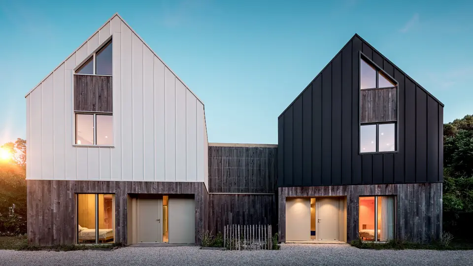 Pohľad z prednej strany na obytný súbor Le Rhune odhaľuje dva vzájomne posunuté objekty s identickým architektonickým výrazom, ktoré sa od seba odlišujú kontrastným farebným riešením v bielej a čiernej. Čisté tvary sedlových striech a súvislý hliníkový plášť PREFALZ dodávajú budovám sochársky charakter a pôsobivo zvýrazňujú precízne línie stojatej drážky. Teplé svetelné akcenty prenikajúce cez veľkorysé presklené plochy vytvárajú výrazný kontrast k minimalistickej architektúre. Odolný a trvácny hliník PREFALZ spája strechu a fasádu do modernej, harmonickej obálky budovy s vysokou estetickou a architektonickou hodnotou.
