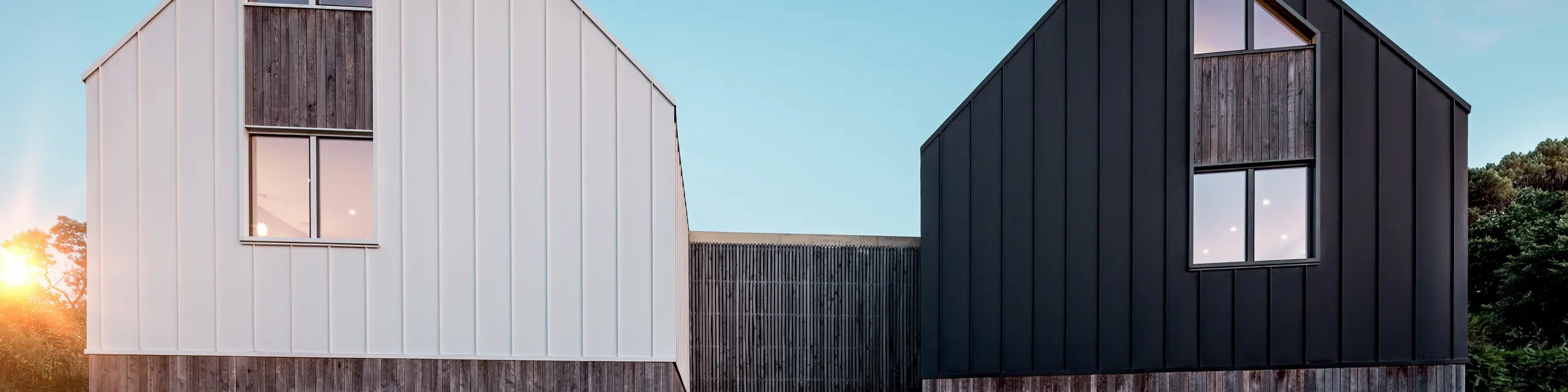 Pohľad z prednej strany na obytný súbor Le Rhune odhaľuje dva vzájomne posunuté objekty s identickým architektonickým výrazom, ktoré sa od seba odlišujú kontrastným farebným riešením v bielej a čiernej. Čisté tvary sedlových striech a súvislý hliníkový plášť PREFALZ dodávajú budovám sochársky charakter a pôsobivo zvýrazňujú precízne línie stojatej drážky. Teplé svetelné akcenty prenikajúce cez veľkorysé presklené plochy vytvárajú výrazný kontrast k minimalistickej architektúre. Odolný a trvácny hliník PREFALZ spája strechu a fasádu do modernej, harmonickej obálky budovy s vysokou estetickou a architektonickou hodnotou.