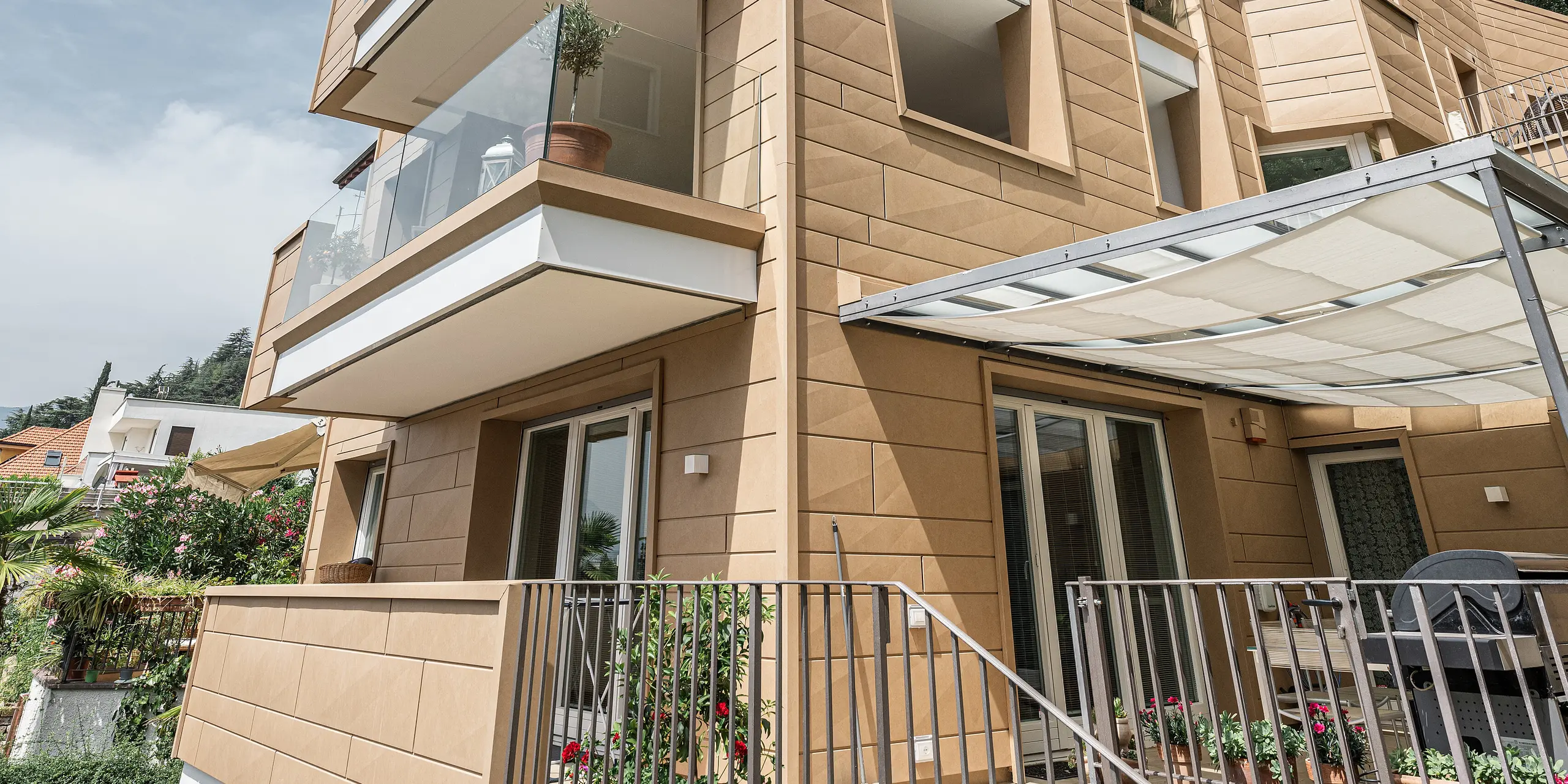 Pohľad hore na fasádu vily Laura, umne navrhnutú pomocou systému PREFA Siding.X v pieskovo hnedej farbe, ktorý spája modernú architektúru a funkčnosť v odolnom hliníkovom obklade. Previsnuté balkóny a veľké okná otvárajú budovu k nebu, zatiaľ čo jemná štruktúra fasádnych panelov a ich prirodzená farebnosť vizuálne uzemňujú budovu a začleňujú ju do stredomorskej krajiny Južného Tirolska.