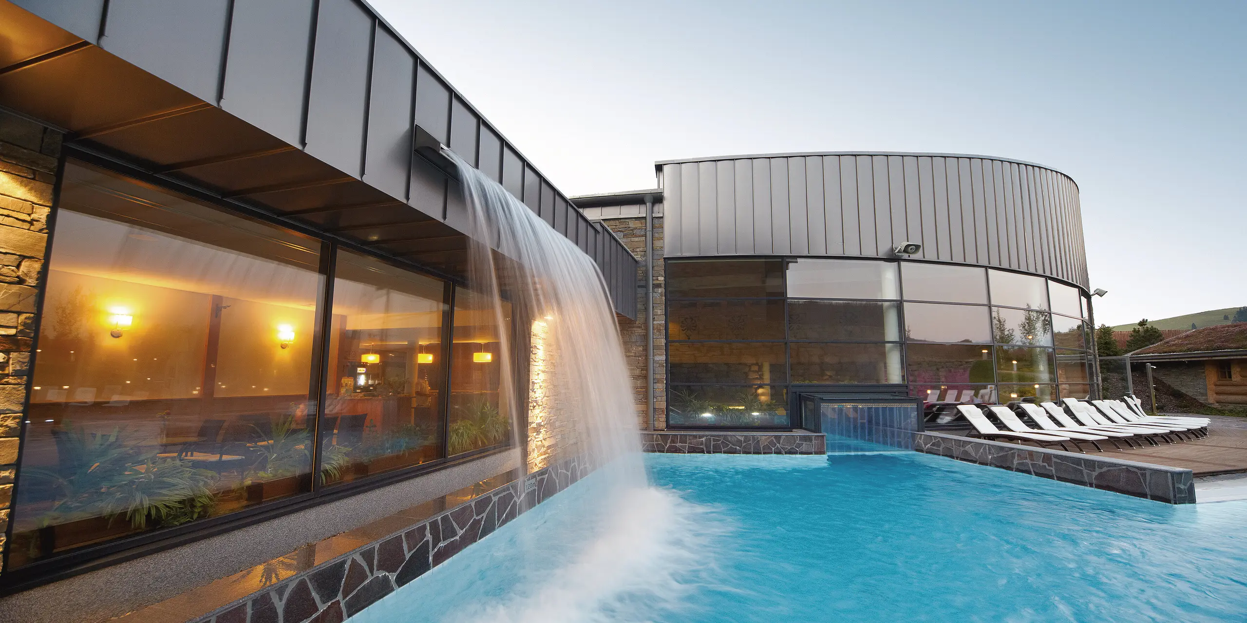 Ein Blick auf den imposanten Wasserfall der Therme Białka zeigt das perfekte Zusammenspiel von Architektur und Natur. Die PREFALZ-Fassade in der Sonderfarbe Delphin ergänzt die organischen Elemente und bildet einen eleganten Kontrast zur Steinmauer und den sanften Wasserbewegungen im Poolbereich.