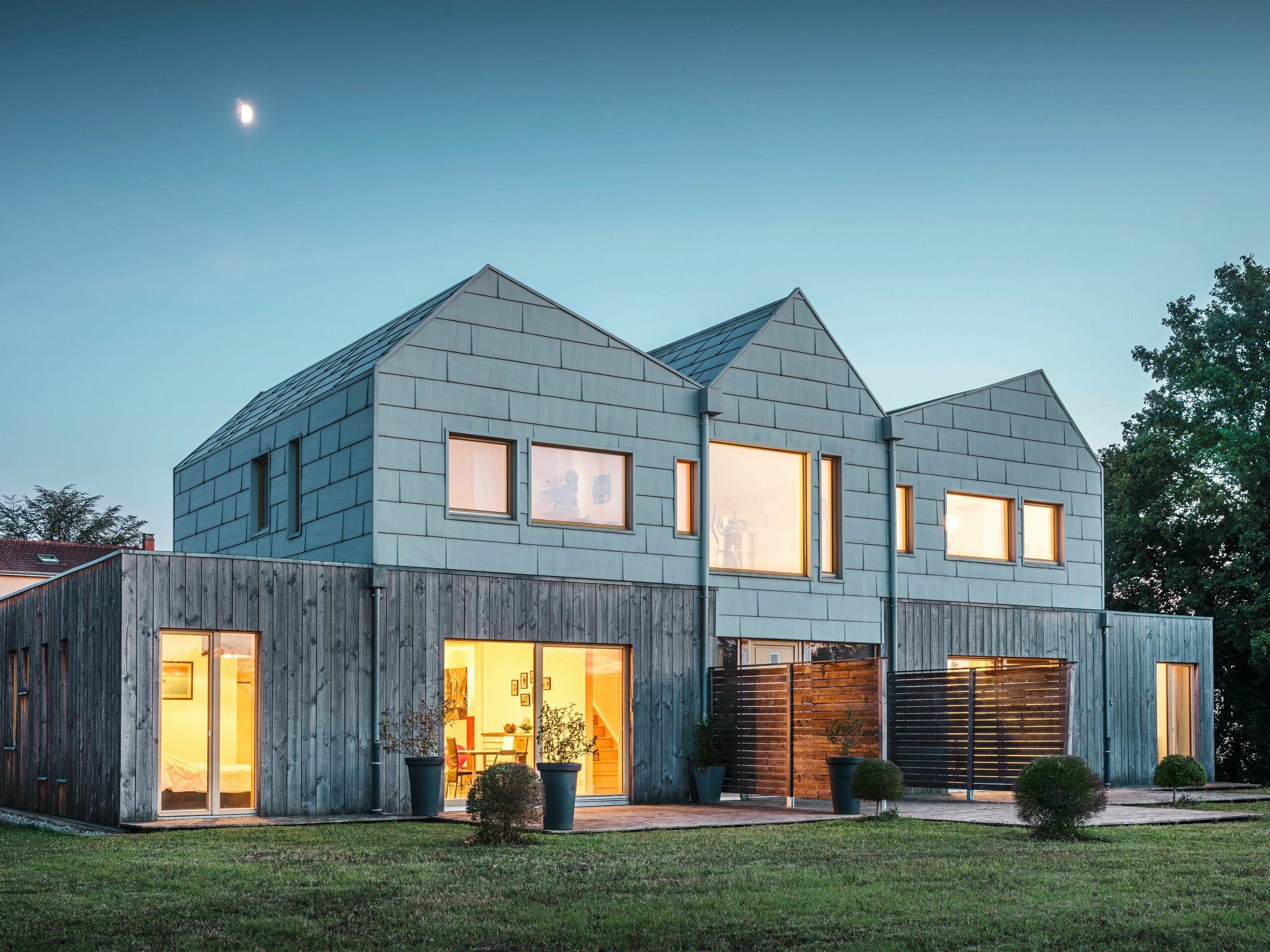 Doppelhaus bei Abenddämmerung mit leuchtenden Innenräumen, die durch große Fenster sichtbar sind. Die Aluminiumpaneele von PREFA in FX.12-Optik fügen sich nahtlos in das moderne Design ein. Das Gebäude ist von einem gepflegten Garten umgeben.