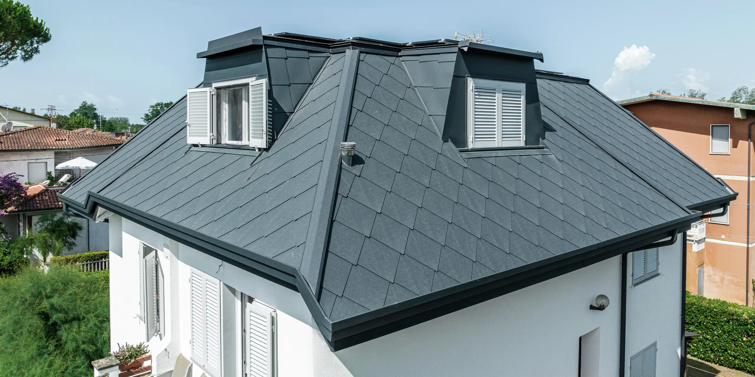 Na tomto detailnom zábere je zreteľne vidieť precízne spracovanie strešnej šablóny PREFA 44 × 44 v kamennej šedej P.10 na rodinnom dome v Montignoso. Vysokokvalitná hliníková strešná krytina poskytuje štýlový, moderný vzhľad a zároveň ponúka maximálnu odolnosť voči extrémnym poveternostným podmienkam. Elegantné vikiere harmonicky splývajú so strechou a vytvárajú ďalší obytný priestor pod robustnou a odolnou strešnou krytinou. Dokonalá súhra dizajnu a funkčnosti.