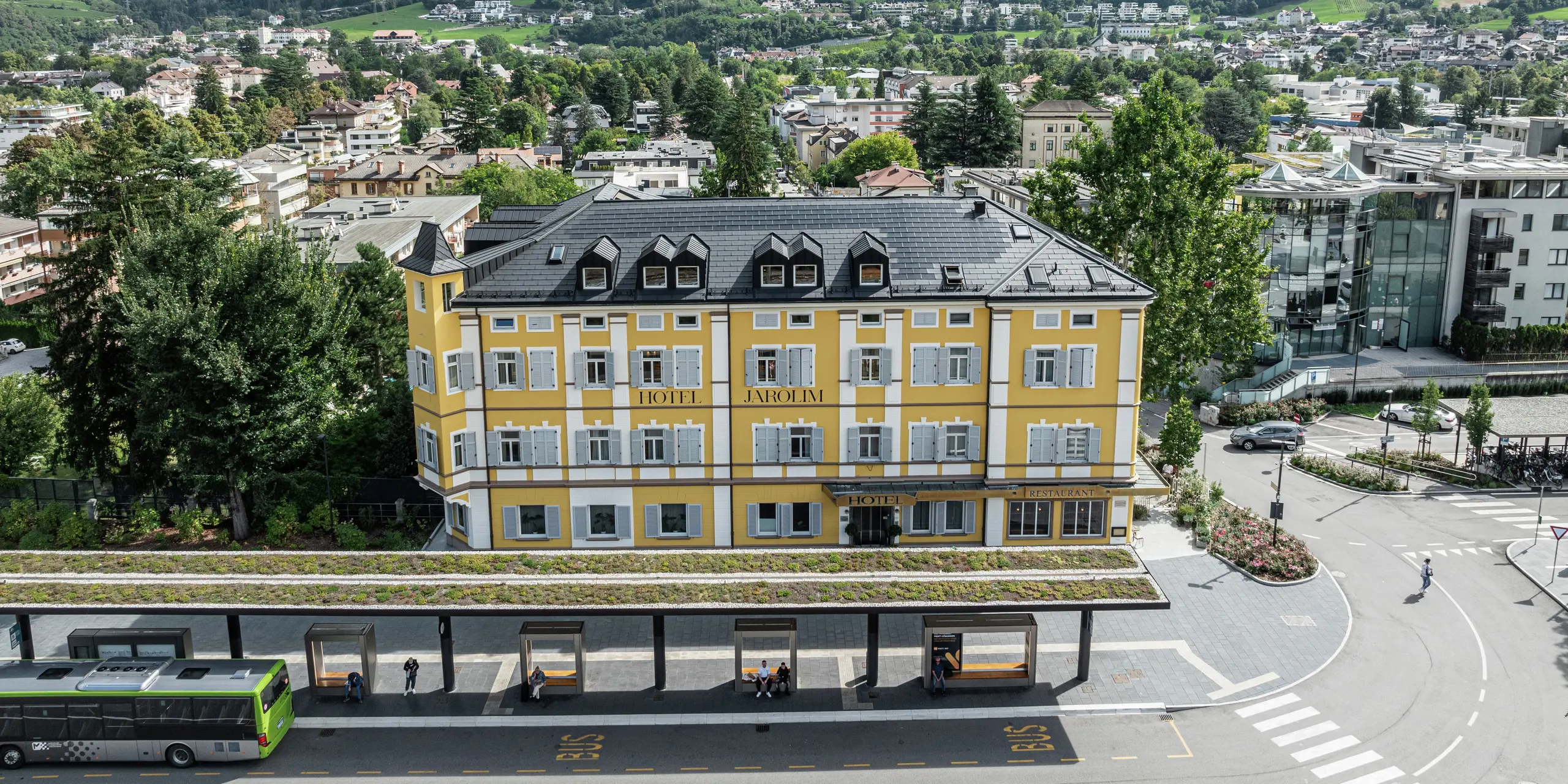 Pohľad zhora na zrekonštruovaný hotel Jarolim pri železničnej stanici v Brixene so strechou z integrovaných solárnych panelov PREFA a strešných panelov R.16. Robustná hliníková strecha presvedčí svojou poveternostne odolnou a ľahkou konštrukciou, ideálnou pre pamiatkovo chránené budovy. Harmonicky integrované fotovoltické moduly zabezpečujú udržateľnú výrobu energie bez narušenia historickej estetiky. Výrazná veža bola navrhnutá s robustnými strešnými šindľami PREFA.