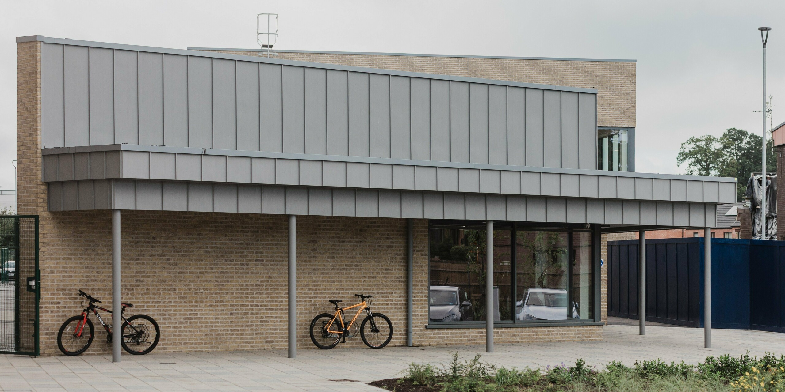 Das Northern Regional College in Ballymena verfügt über einen modernen Fahrradstellplatz mit großzügigem Vordach aus PREFALZ Aluminium in der Farbe Patinagrau. Die vertikal strukturierten Stehfalzbahnen ziehen sich präzise über die Dachkante und bilden einen klaren Blickfang über der Klinkerfassade. Schlanke Stützen, helle Klinkerwände, abgestellte Fahrräder und der offene Vorplatz unterstreichen den nutzerfreundlichen Charakter des Campus. PREFA Aluminium überzeugt durch Langlebigkeit, Witterungsbeständigkeit und eine ruhige, hochwertige Optik im täglichen Betrieb.