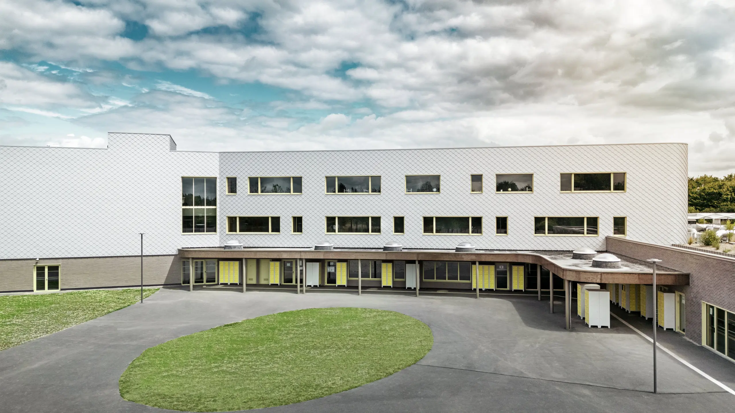 Zadná strana školy Collège Léontine Dolivet s veľkým školským dvorom a čistou architektúrou. Fasáda je tvorená striebornými kovovými fasádnymi šablónami PREFA, ktoré jej dodávajú moderný, elegantný vzhľad. Niekoľko radov okien a žlté dvere oživujú štruktúru, zatiaľ čo asfaltové nádvorie s centrálnou zelenou plochou ponúka veľa priestoru na prestávky. Odolná hliníková fasáda kombinuje ľahkosť, odolnosť voči poveternostným vplyvom a dizajn – funkčný a estetický pohľad v Cesson-Sévigné neďaleko Rennes.