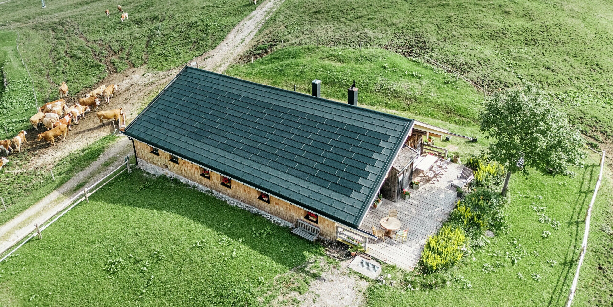 Pohľad z vtáčej perspektívy na brezovú chatu na Walleralme, Bayrischzell, s novým strešným plášťom zo strešných panelov PREFA R.16 vo farbe P.10 čierna a integrovaných solárnych panelov PREFA. Tradičná drevená chata s útulnou terasou dokonale zapadá do zelenej horskej krajiny. Na okolitých lúkach sa pokojne pasú kravy, zatiaľ čo inovatívny strešný systém podporuje udržateľnú výrobu energie a zároveň poskytuje robustnú ochranu pred poveternostnými vplyvmi.