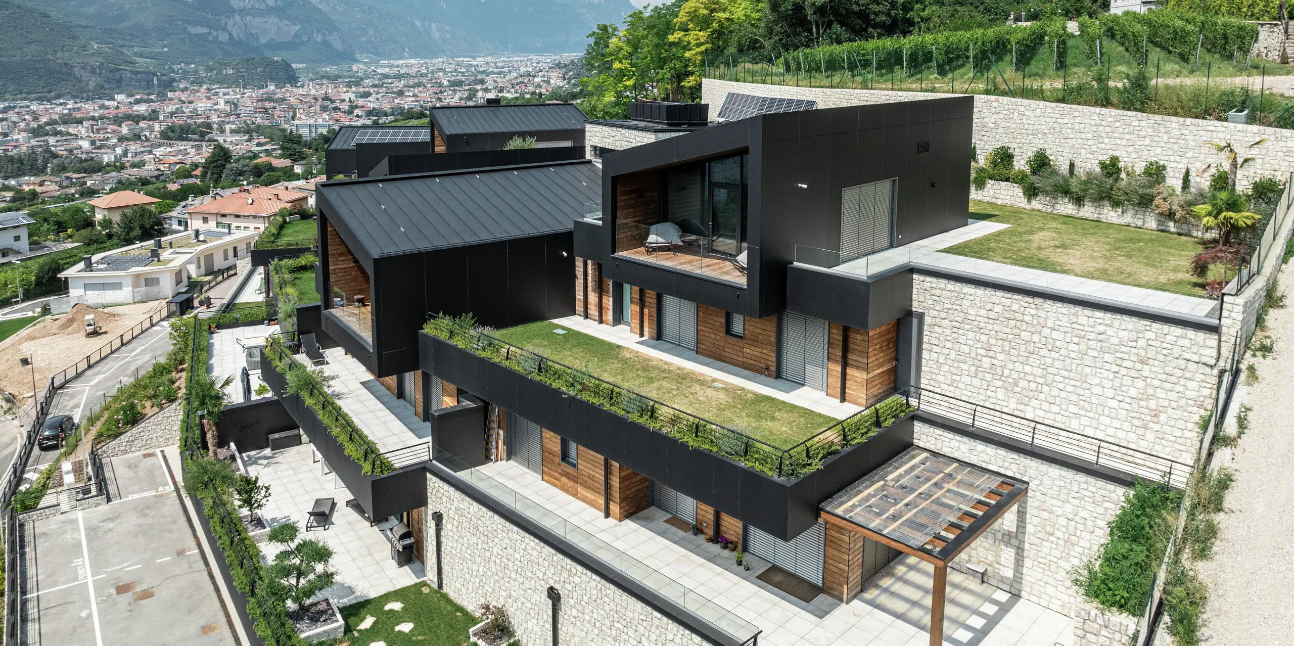 Exkluzívny rezidenčný komplex s panoramatickým výhľadom na Trento: Moderná architektúra harmonicky zapadá do svahovitej polohy a kombinuje vysoko kvalitné materiály, ako je prírodný kameň, drevo a hliník. Fasáda z hliníkových kompozitných dosiek PREFA v čiernej farbe P.10 zaisťuje elegantný a trvalý vzhľad, zatiaľ čo strecha PREFALZ v čiernej farbe P.10 poskytuje optimálnu ochranu pred poveternostnými vplyvmi. Zelené plochy a priestranné terasy podčiarkujú udržateľný koncept bývania a vytvárajú dokonalé spojenie prírody a moderného dizajnu.