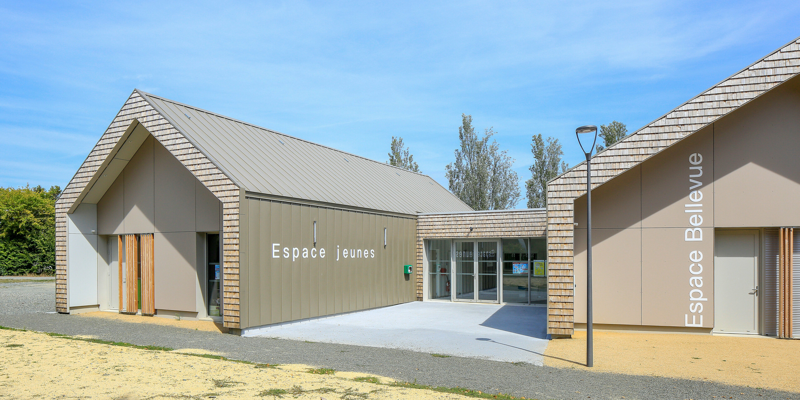 Salles Espace Bellevue v Mûrs-Erigné, Francúzsko, s nápismi „Espace Jeunes“ a „Espace Bellevue“ pred halou podujatia. Strešný a fasádny systém PREFALZ v bronzovej farbe zdobí veľkú časť moderného mládežníckeho centra. Hliníkové opláštenie na stojatú drážku od spoločnosti PREFA odolné voči poveternostným vplyvom ponúka trvanlivé a moderné riešenie pre stavebné projekty. Kombinácia hliníkového obkladu a prírodného vzhľadu dreva harmonicky zapadá do okolia.