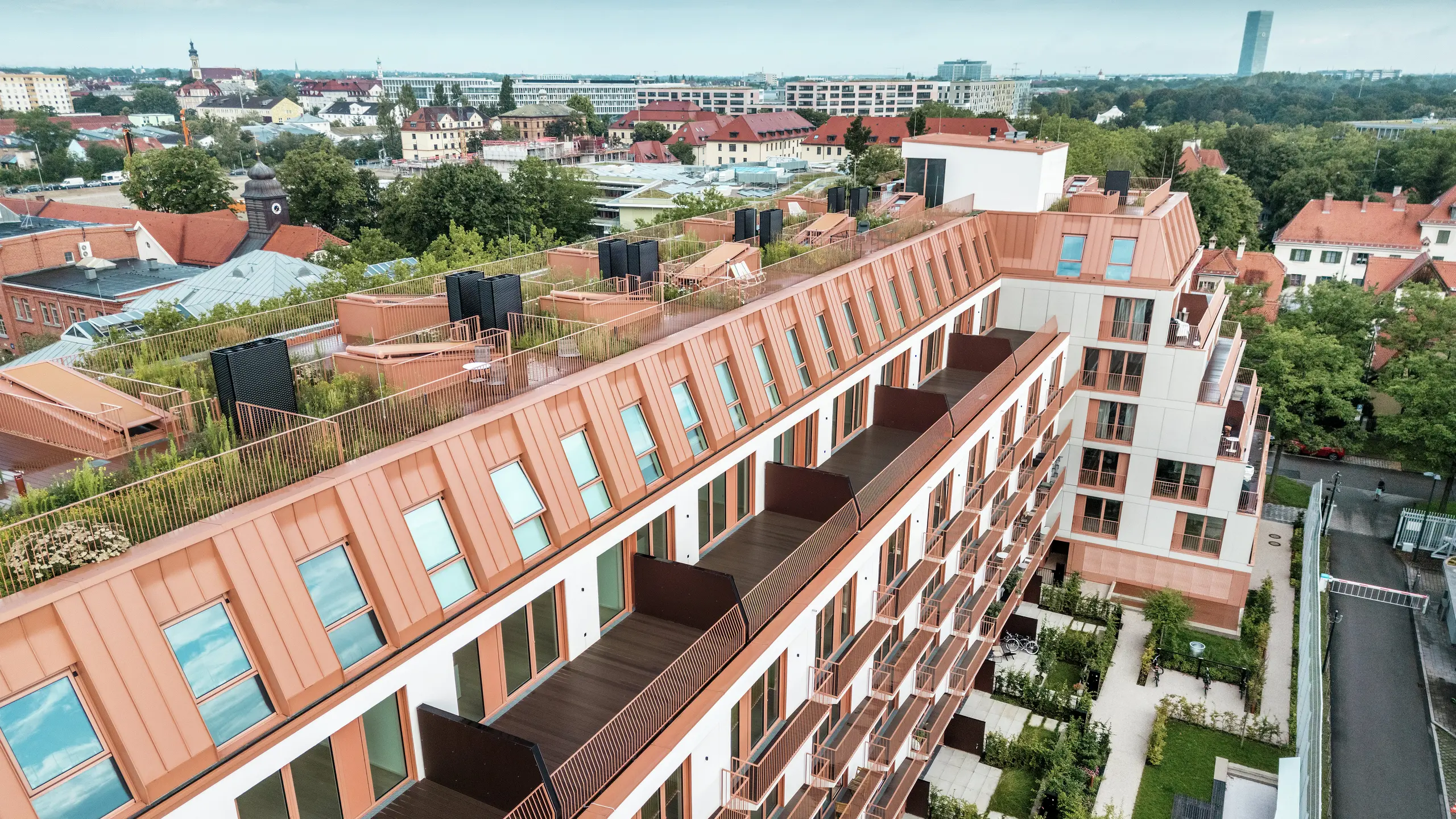 Letecký pohľad na obytný projekt Van B v Mníchove s výraznou hliníkovou strechou PREFALZ v špeciálnej farbe IGP Orange HDP od spoločnosti PREFA. Priestranné balkóny, zelené strešné terasy a precízny dizajn fasády zdôrazňujú premyslený architektonický koncept. Ideálne riešenie pre udržateľný rozvoj mesta a mestské bývanie s vysokou kvalitou života a estetickou príťažlivosťou.