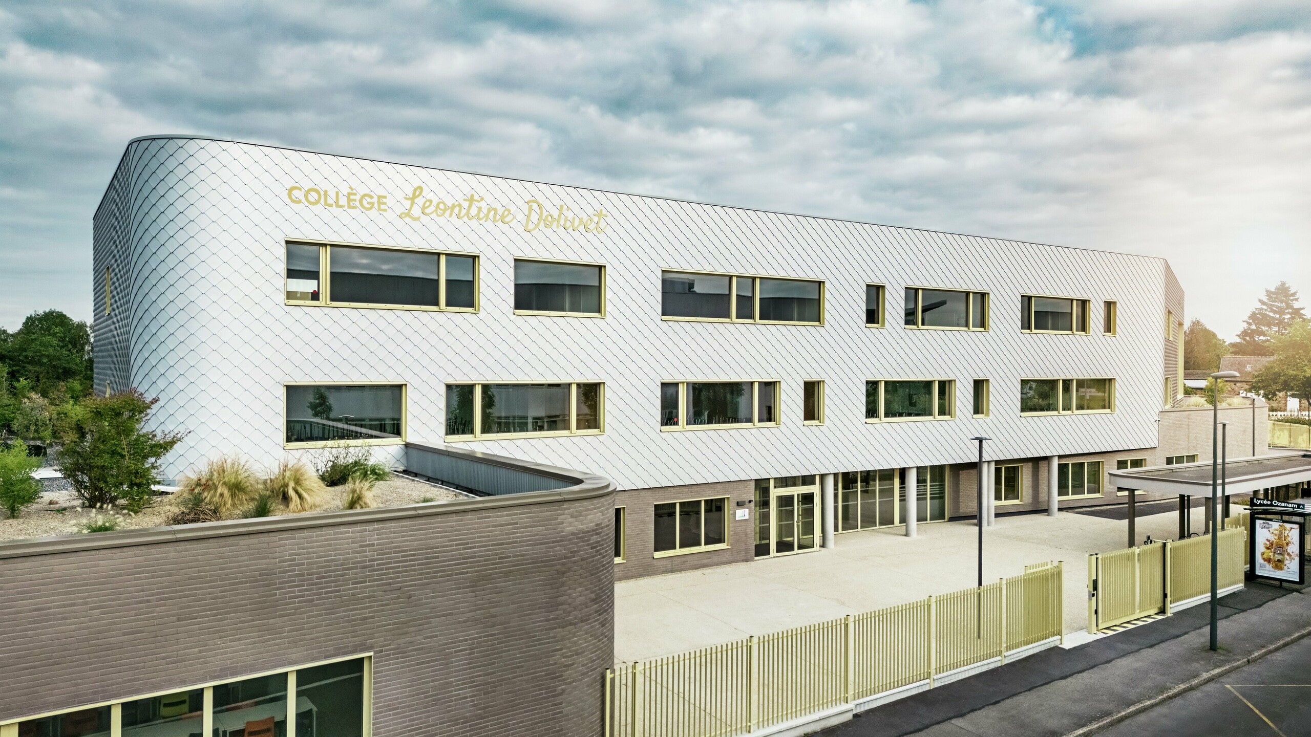 Pohľad na fasádu školy Collège Léontine Dolivet v Cesson-Sévigné s modernou hliníkovou fasádou z PREFA fasádnych šablón v striebornom metalickom vyhotovení. Meniaci sa povrch dodáva budove ľahkosť, zatiaľ čo čisté línie a veľké okná zaisťujú transparentnosť. Žlté okenné rámy dodávajú budove živý akcent. V popredí je vidieť svetlé predsieň, plot a susednú ulicu. Trvanlivé fasádne riešenie PREFA kombinuje dizajn, odolnosť voči poveternostným vplyvom a udržateľnosť – architektonický pohľad upútavajúci elegantným dizajnom.