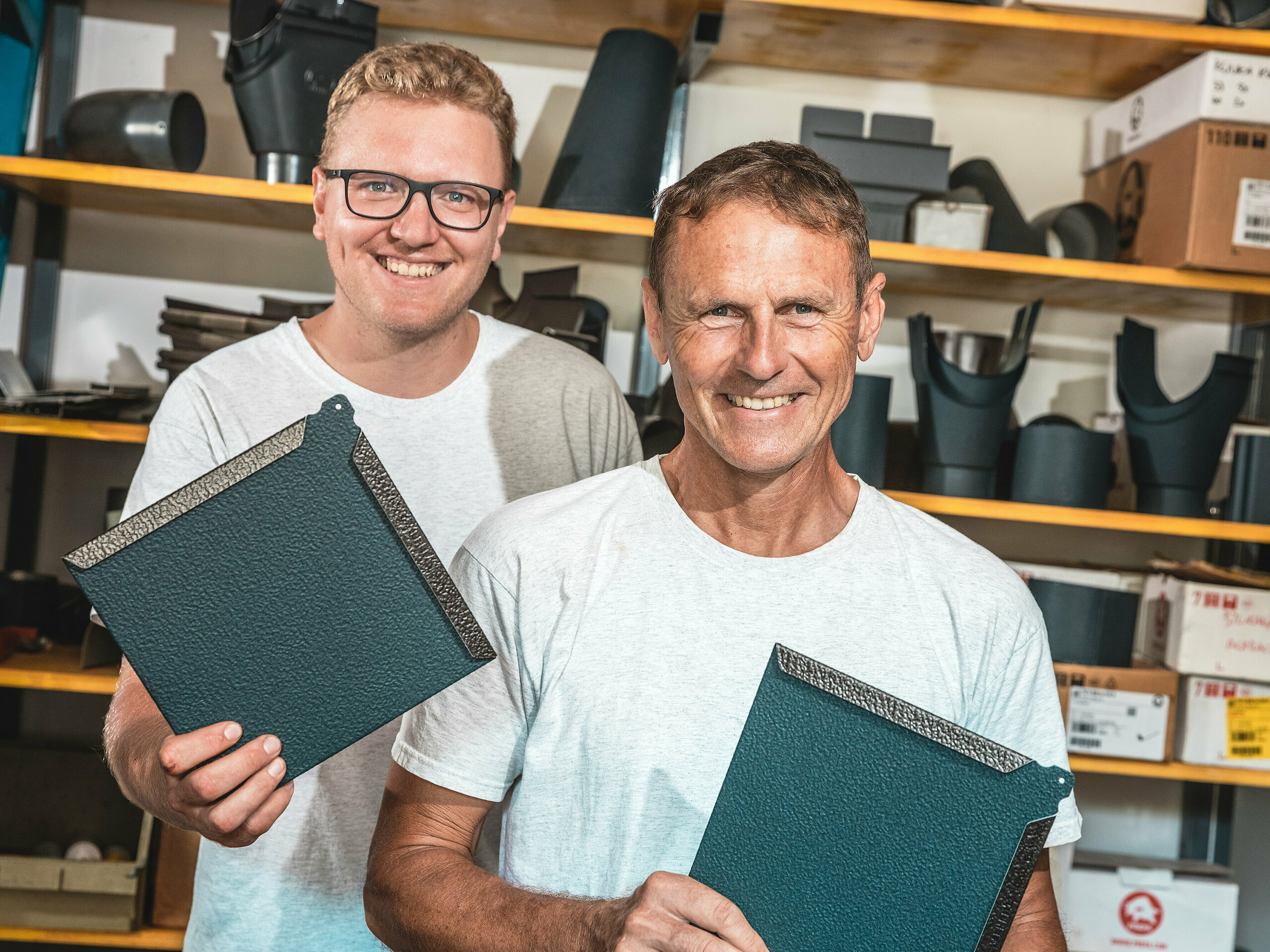 V dielni dvaja remeselníci prezentujú štvorcové hliníkové strešné šablóny PREFA v farbe P.10 antracit. Muž v popredí je slovinský klampiarsky majster Janez Kralj. Za ním stojí mladý spolupracovník z jeho tímu. V pozadí sú vidieť regály s komponentmi a príslušenstvom pre strešné a fasádne systémy. Jemne štruktúrovaný povrch hliníkových šablón PREFA svedčí o vysokej kvalite materiálu, dlhej životnosti a precíznom spracovaní. Sú ideálne pre náročné architektonické projekty, ako sú tiny houses, kde sa vyžaduje odolnosť, nízka hmotnosť a moderný dizajn.