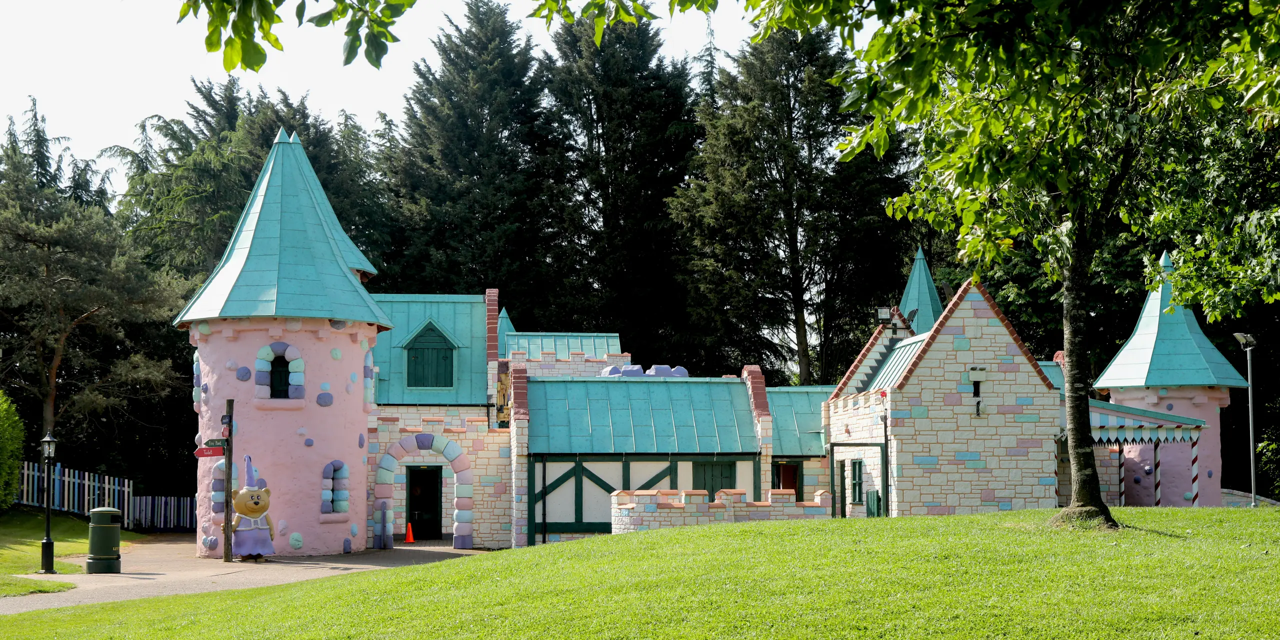 Weitläufige Ansicht der märchenhaften Schlossanlage im Sundown Adventureland, eingebettet in eine grüne Parklandschaft mit altem Baumbestand als natürlicher Rahmen. Verspielte Rundtürme, filigrane Giebel und unterschiedlich geneigte Dächer bilden spannende Eyecatcher. Die PREFALZ Aluminiumdächer in P.10 Patinagrün setzen prägnante Akzente und betonen die komplexe Architektur mit klaren Stehfalzen. Dank hoher Formbarkeit gelingen selbst anspruchsvolle Details, während das PREFA System durch Langlebigkeit, Korrosionsbeständigkeit und zuverlässigen Wetterschutz überzeugt.