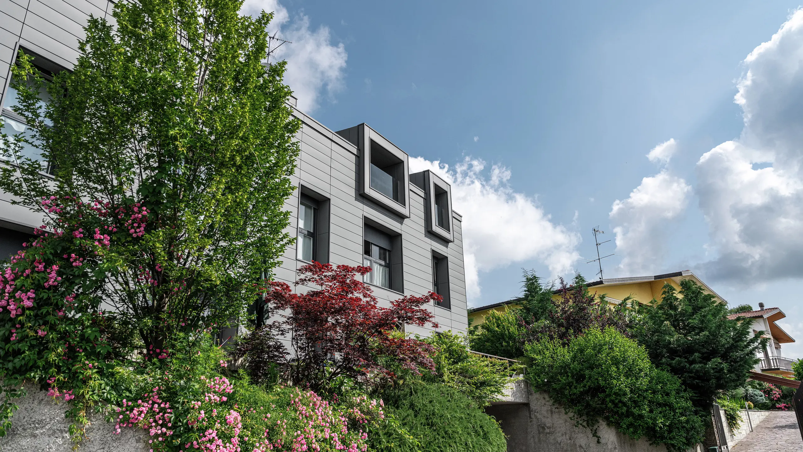 Moderný obytný komplex v talianskom meste Faloppio so štýlovou fasádou z lamelových dosiek PREFA Siding. Vyčnievajúce okná sú jedinečnou architektonickou ozdobou, zatiaľ čo okolité rastliny a kvety zdôrazňujú prirodzenú krásu pozemku. Odolný hliníkový obklad PREFA poskytuje estetické a funkčné riešenie za každého počasia.