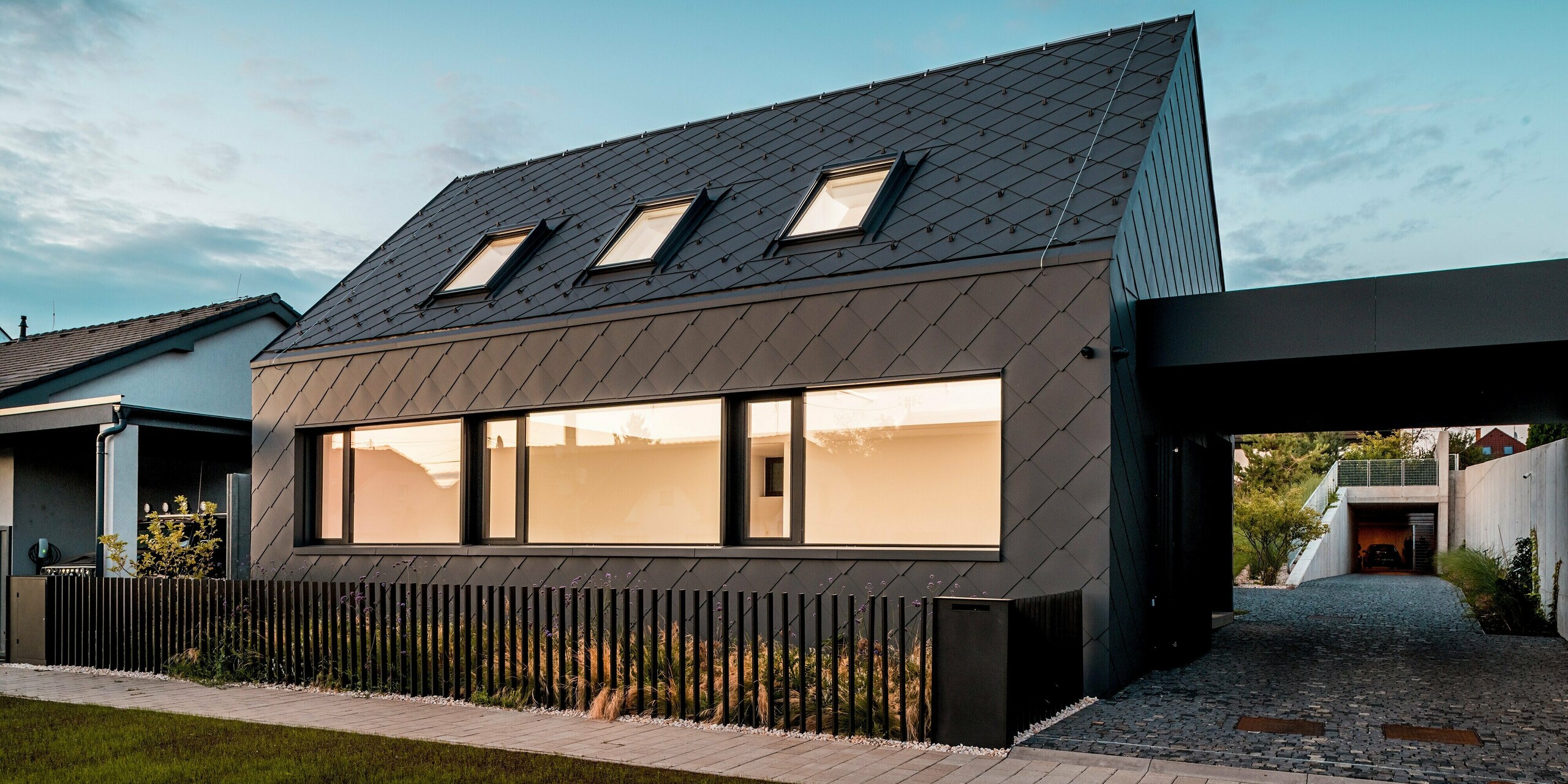 Na obrázku je moderný rodinný dom v Trnave za súmraku, kompletne obložený strešnými a fasádnymi šablónami PREFA 44 × 44 v odtieni P.10 tmavošedá. Osvetlené okná vytvárajú príjemný kontrast s tmavým, rovnomerne štruktúrovaným hliníkovým plášťom. Presne položený šablónový vzor zdôrazňuje čisté línie budovy a podčiarkuje vysokú kvalitu a odolnosť použitých materiálov. Odolné hliníkové produkty PREFA poskytujú optimálnu ochranu pred poveternostnými vplyvmi a dodávajú budove výrazný, nadčasový vzhľad.