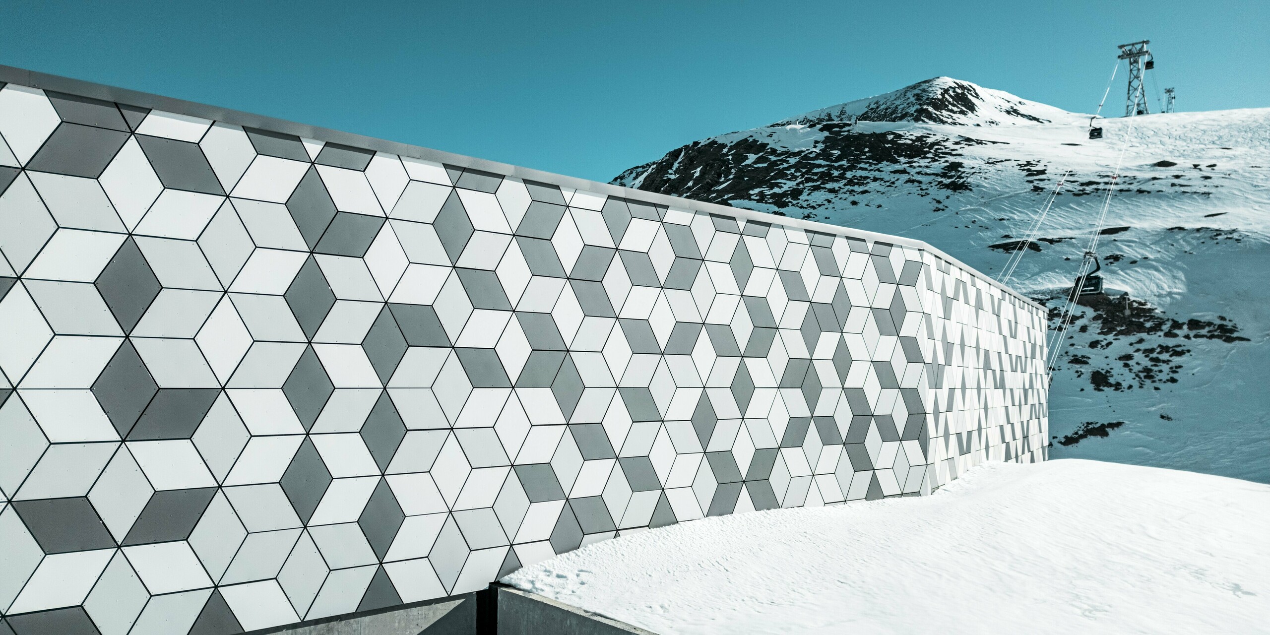 Fasáda stanice lanovky v Les Deux Alpes je majstrovským dielom modernej architektúry. Pozostáva z hliníkových kompozitných panelov PREFABOND usporiadaných do pútavého kosoštvorcového vzoru bielej a sivej farby. Geometrický dizajn harmonicky zapadá do zasneženej horskej krajiny a vďaka robustným materiálom odolným voči poveternostným vplyvom poskytuje dlhodobú ochranu pred extrémnymi poveternostnými podmienkami. Pohľad za fasádou zobrazuje lanovku, ktorá premáva na horu.