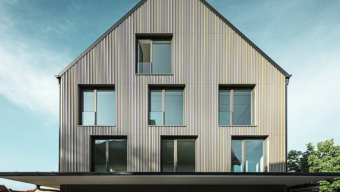 Na obrázku je zobrazená moderná budova s fasádnym obkladom z materiálu Prefalz vo farbe bronzová a šikmou strechou, ktorá je tiež pokrytá materiálom Prefalz v čiernošedej farbe. Veľká sklenená fasáda na prízemí je orámovaná stromami a vytvára príjemnú atmosféru pre reštauračné priestory pod ňou. Kombinácia kvalitných materiálov Prefalz a modernej architektúry dodáva budove elegantný a súčasný vzhľad.