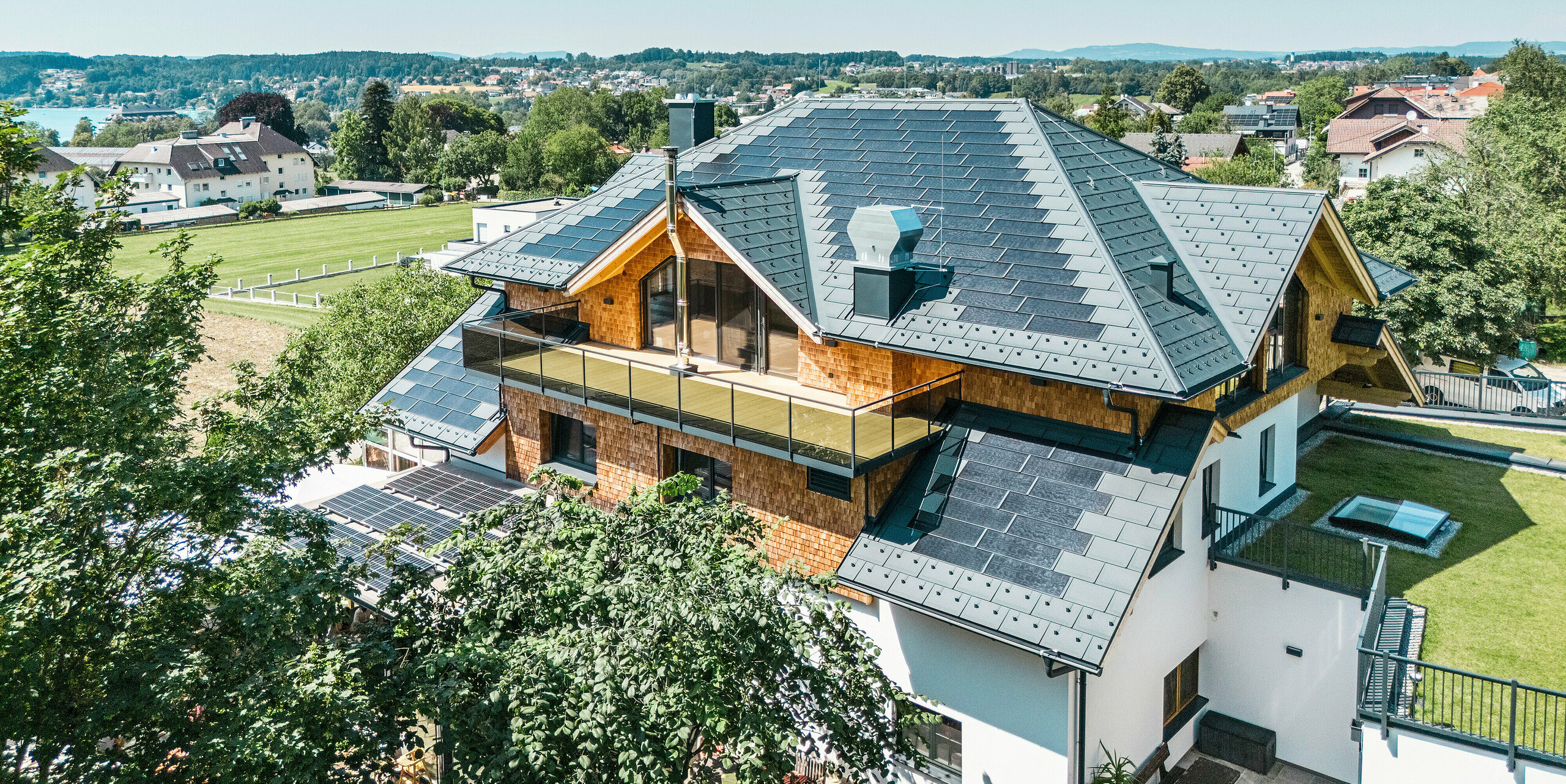 Pohľad z vtáčej perspektívy na Zenz'n Stub'n v Schörflingu pri jazere Attersee s novo zrekonštruovanou strechou z integrovaných solárnych panelov PREFA a strešných panelov R.16 vo farbe P.10 antracitová. Kvalitná plechová strecha spája moderný dizajn s udržateľnou výrobou energie. Priestranný balkón a harmonicky integrované odvodnenie strechy podčiarkujú štýlový vzhľad budovy.