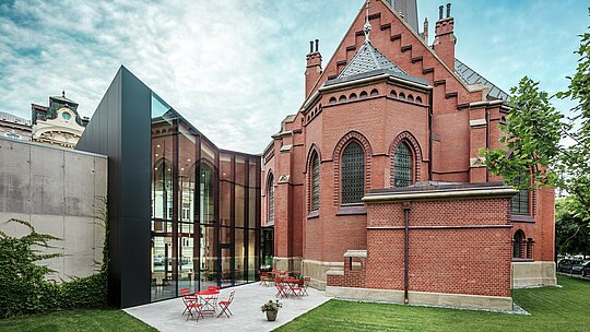 Obrázok ukazuje impozantnú novostavbu, ktorá spája historický Červený kostol s modernou vedeckou knižnicou v Olomouci. Perspektíva ukazuje zadnú časť budovy s príjemnou terasou. Elegantná fasáda z čiernych kompozitných dosiek PREFABOND Aluminium dodáva budove výrazný moderný akcent a štýlovo kontrastuje s červenými tehlami kostola. Vysoká presnosť montáže a použitie odolných materiálov, ako je hliník, zaručujú harmonické spojenie tradície a inovácie. Tieto pokrokové dosky sú nielen esteticky príťažlivé, ale aj mimoriadne funkčné a odolné, čím sa projekt stáva majstrovským dielom modernej architektúry.