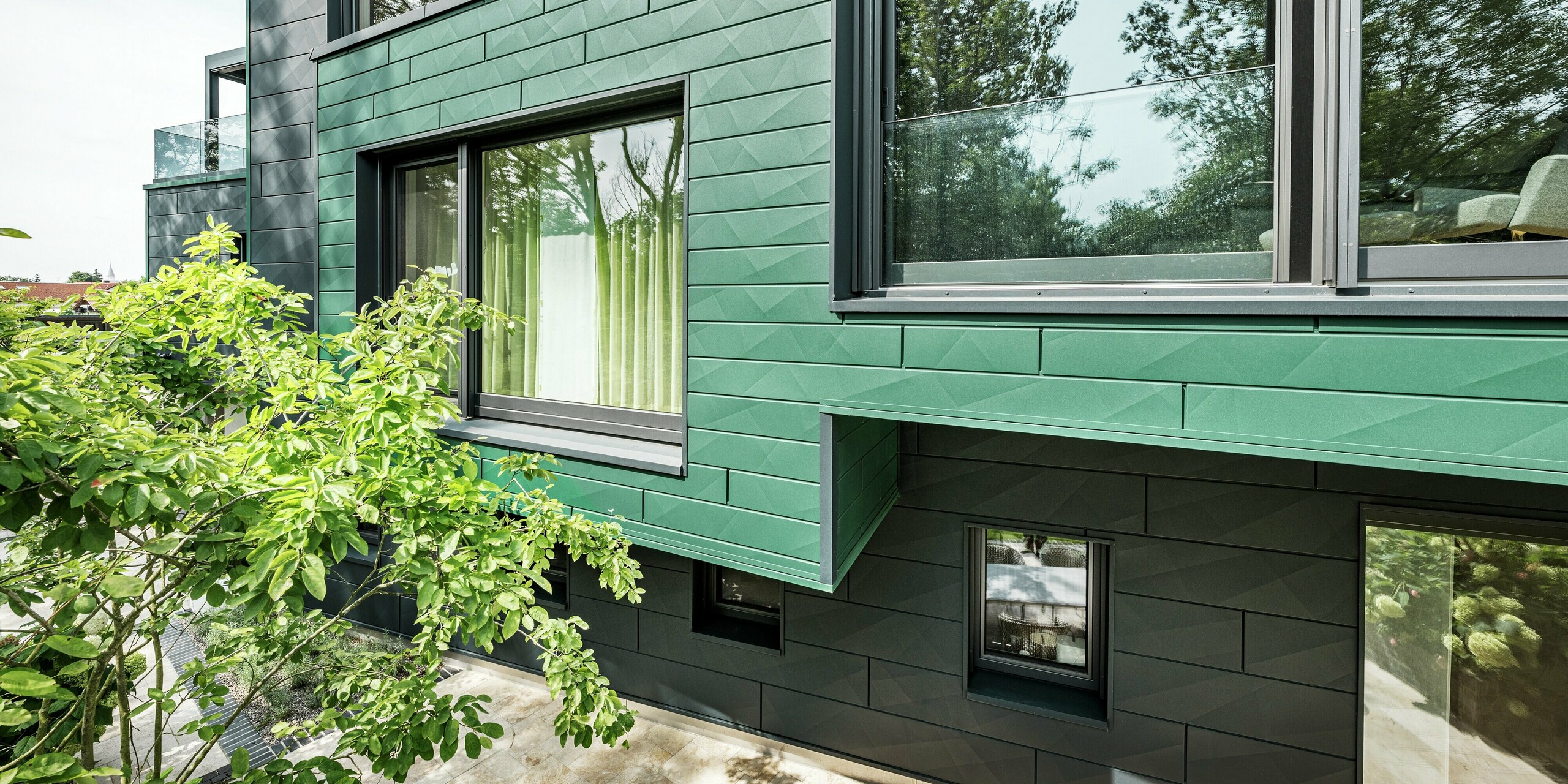 Das Bild zeigt die markante Fassade eines modernen Einfamilienhauses in Türkheim, das mit PREFA Siding X-Paneelen in den Farben Moosgrün und Anthrazit verkleidet wurde. Die Längs- und Querkantungen der Paneele schaffen eine lebendige Struktur und verleihen dem Gebäude eine dynamische Optik. Die Aluminiumfassade ist witterungsbeständig und pflegeleicht und schützt das Haus somit sowohl optisch als auch funktional.